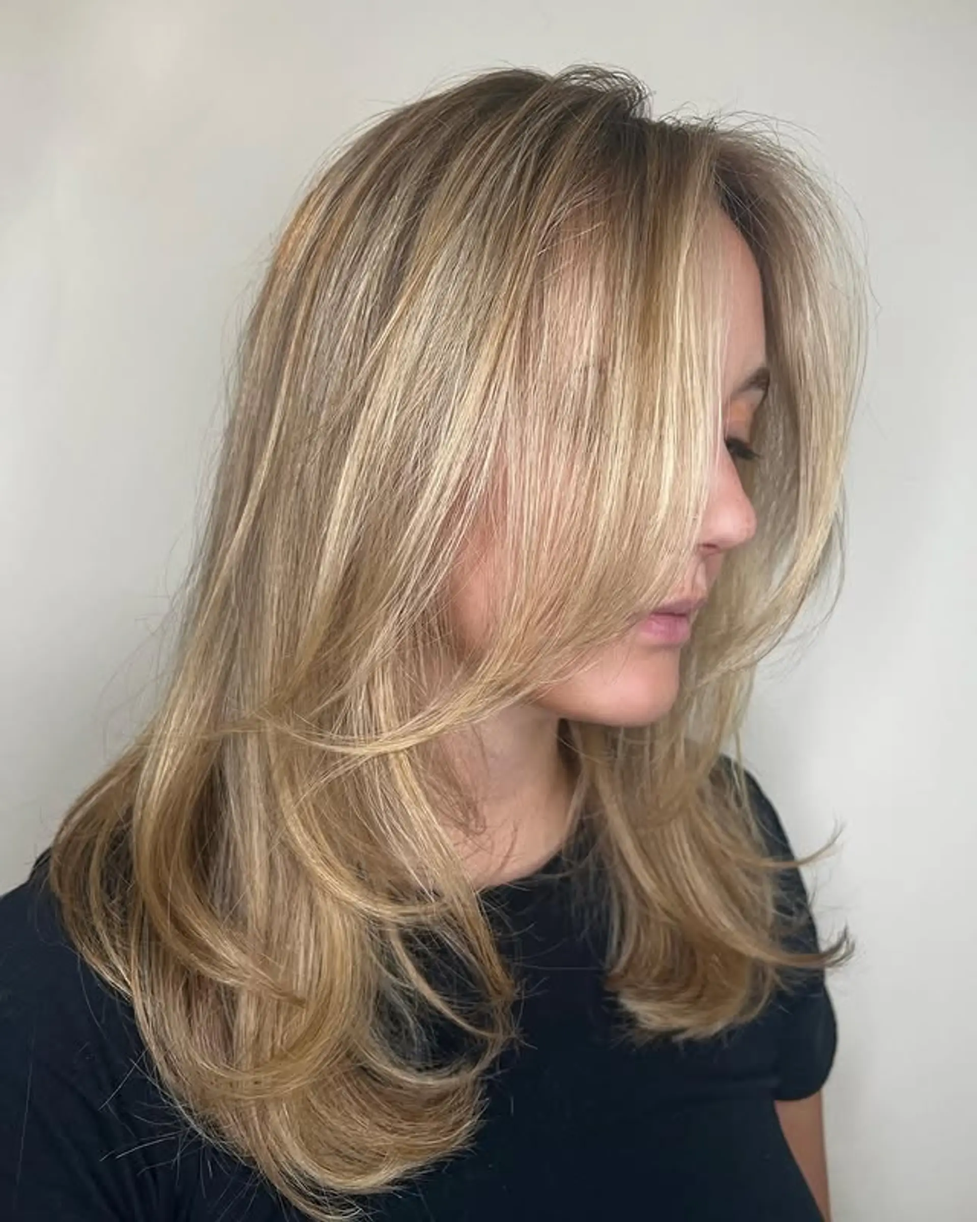 A medium length layered hairstyle featuring face-framing curtain bangs and a bronde color. 1