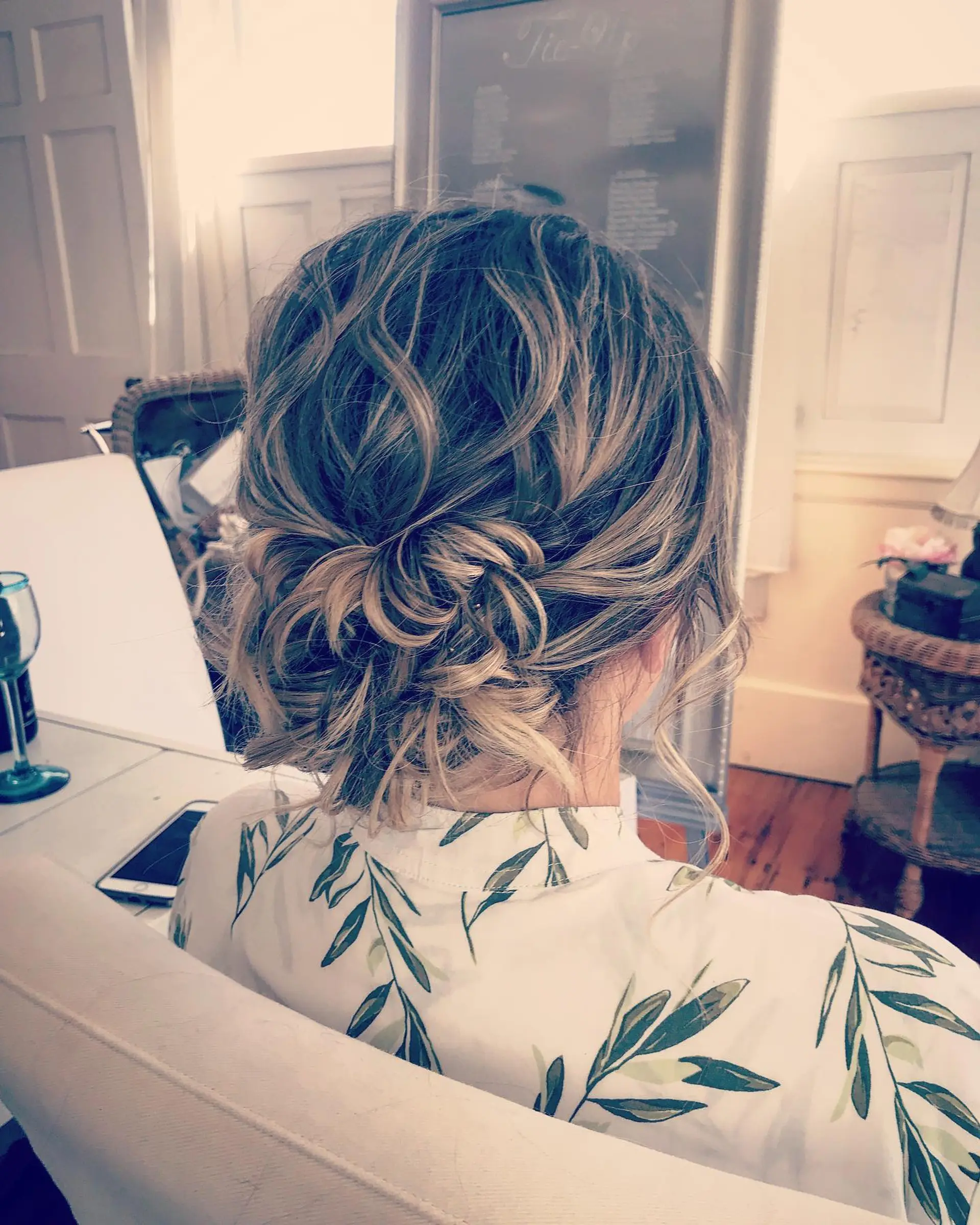 A loose, textured low bun updo featuring soft waves, dimensional blonde highlights, and romantic tendrils. 1