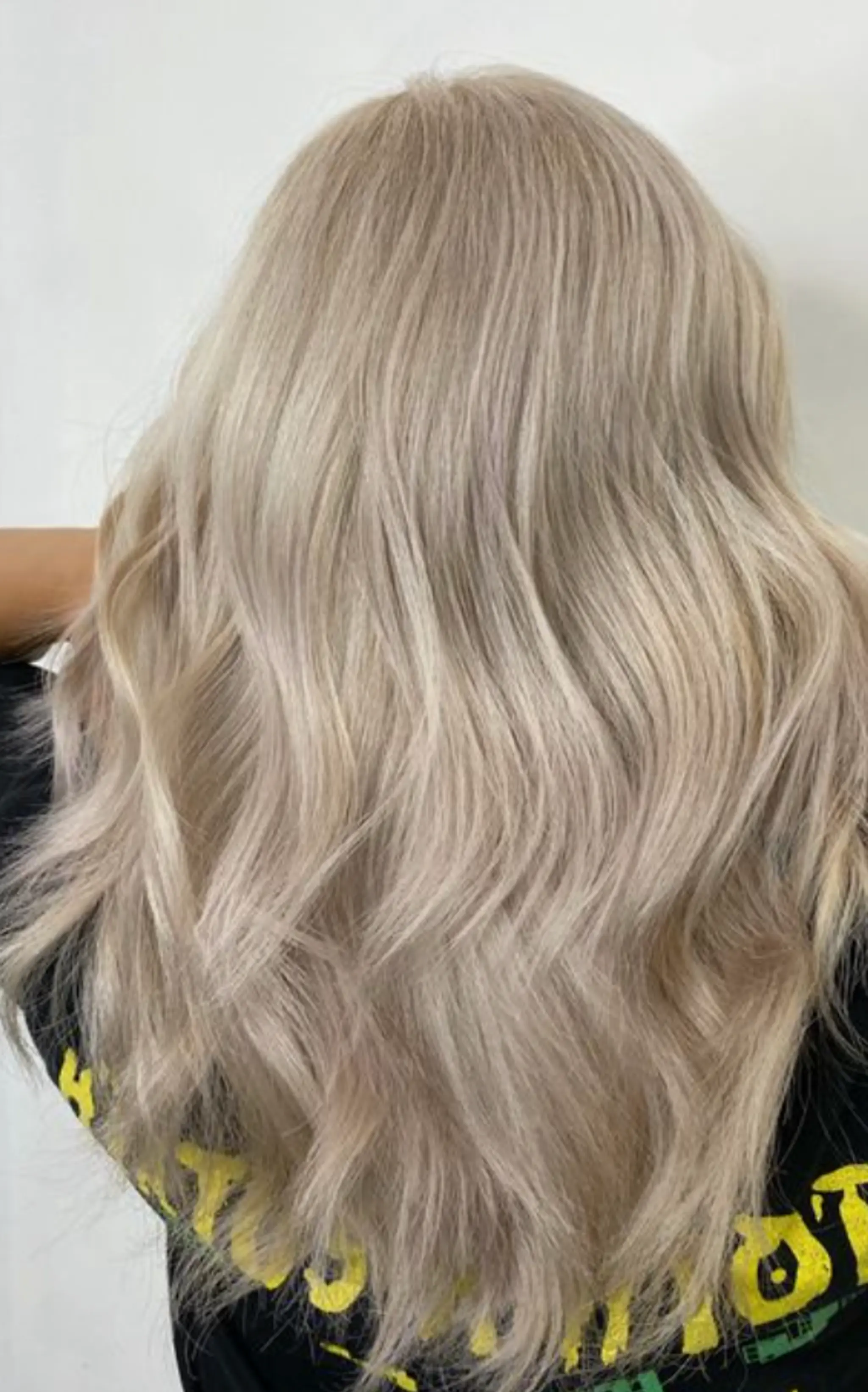 The hairstyle features long, wavy hair with a platinum blonde color. The waves are loose and natural, adding texture and movement throughout the length. The hair is silky and smooth, creating a luxurious and polished look. There are subtle layers that enhance the waves, giving the overall style a voluminous and airy feel.
