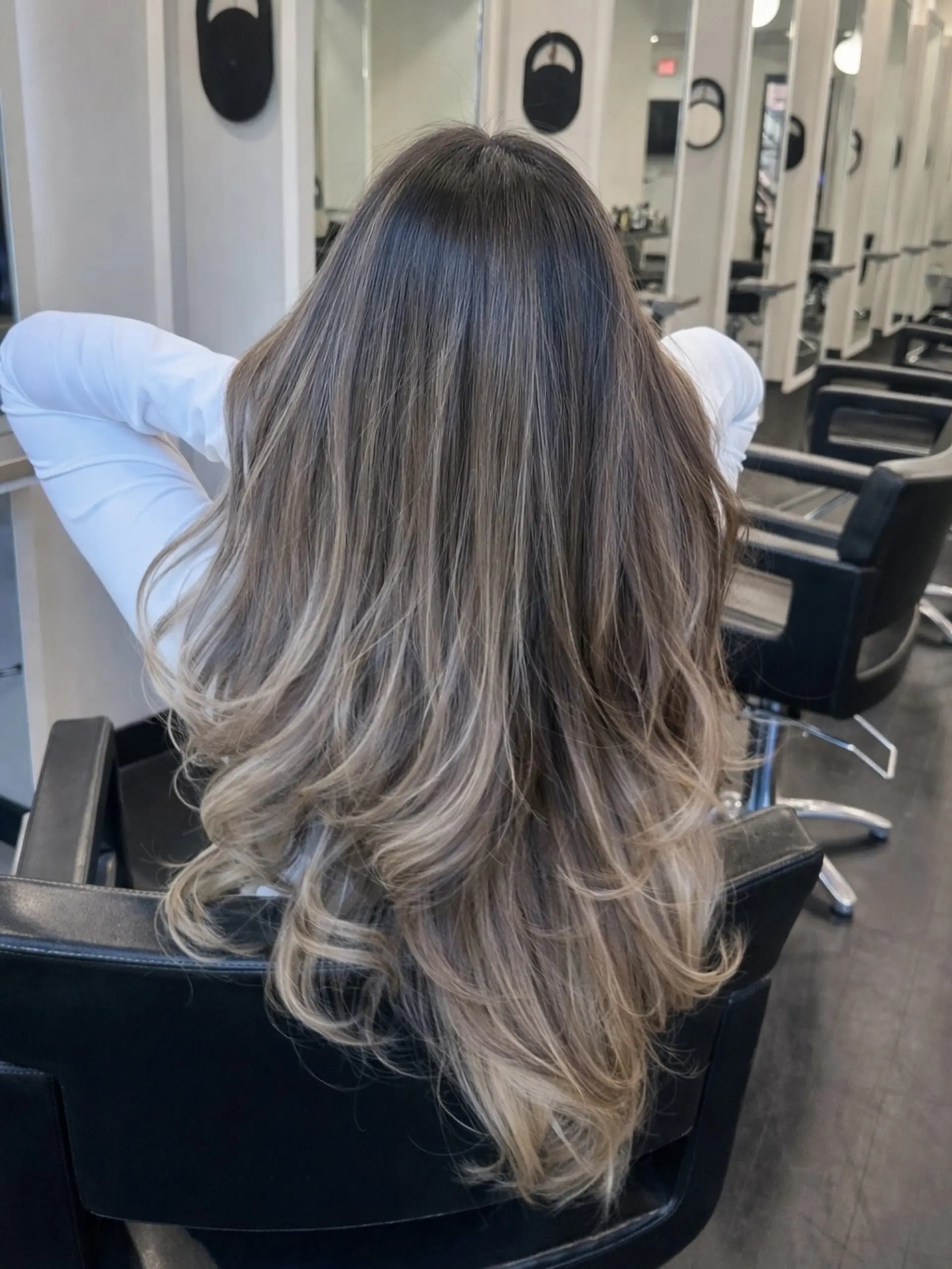 Long dark brown hair seamlessly blended into soft beige blonde balayage highlights, featuring long cascading layers and bouncy curled ends. 1