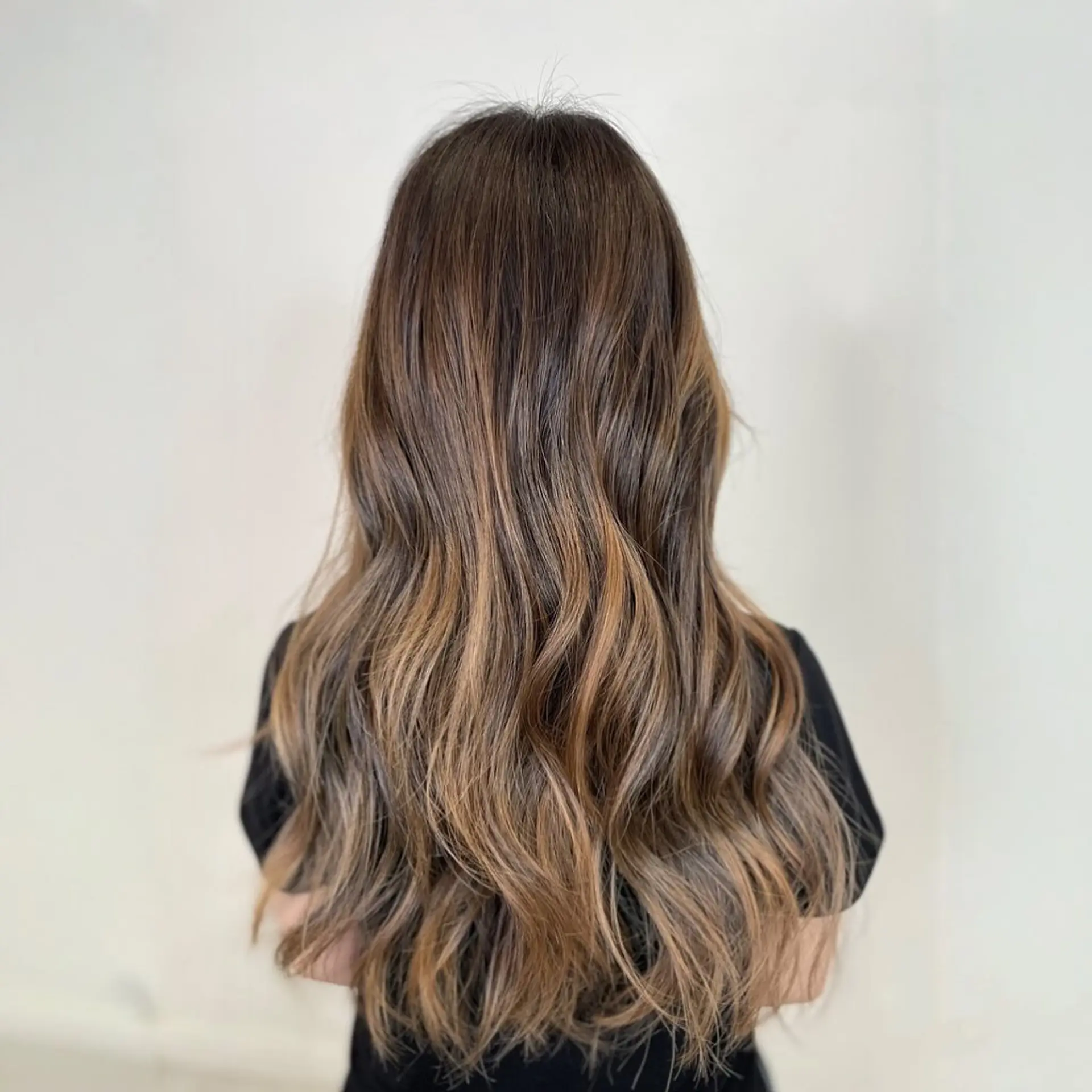 Long dark brown hair seamlessly blended with warm caramel balayage highlights, styled in soft, loose waves. 1