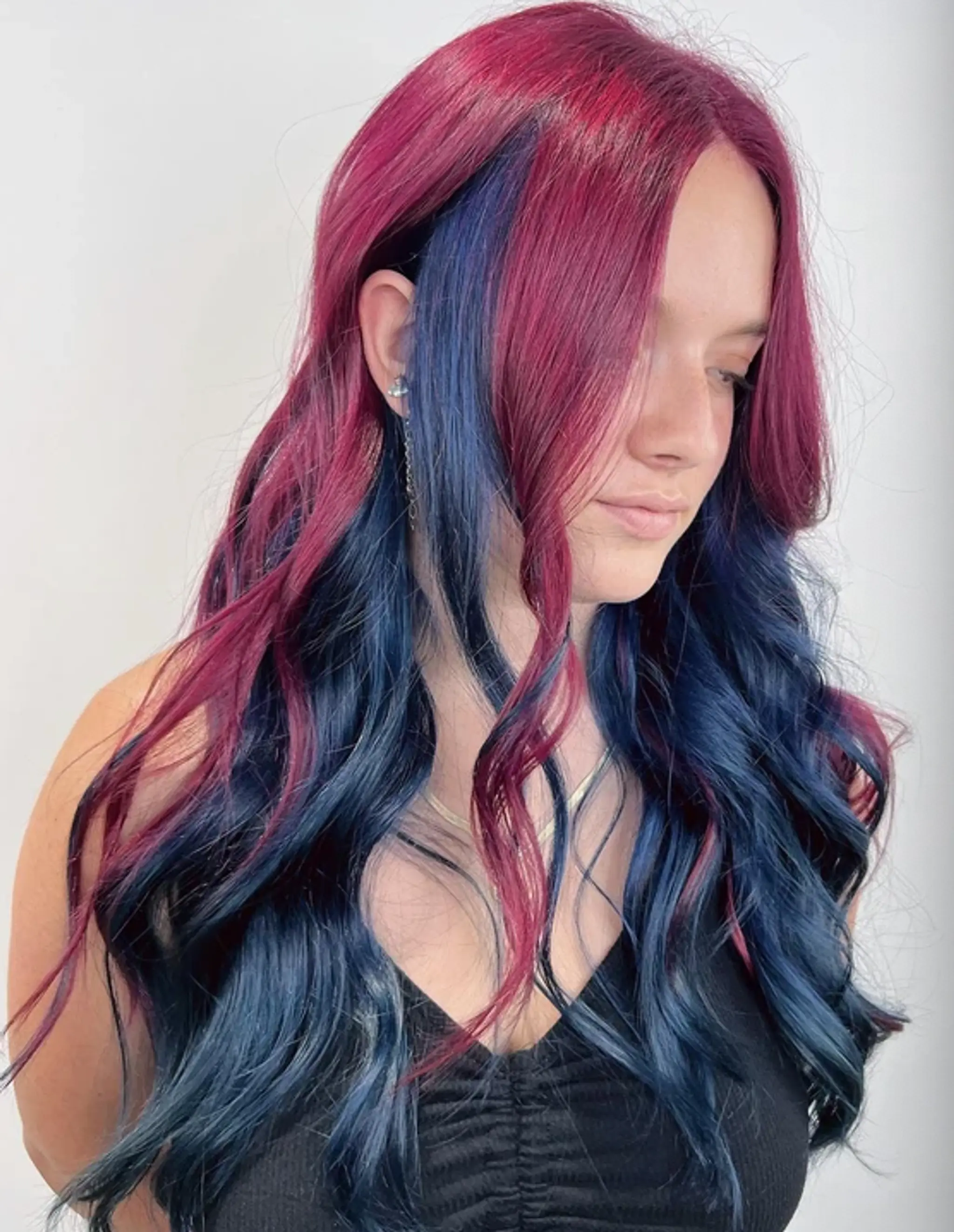 This vibrant hairstyle features long, wavy hair with a striking half-and-half color design, transitioning from rich magenta at the top to deep navy blue at the ends. Loose waves add volume and texture, while face-framing layers soften the look.