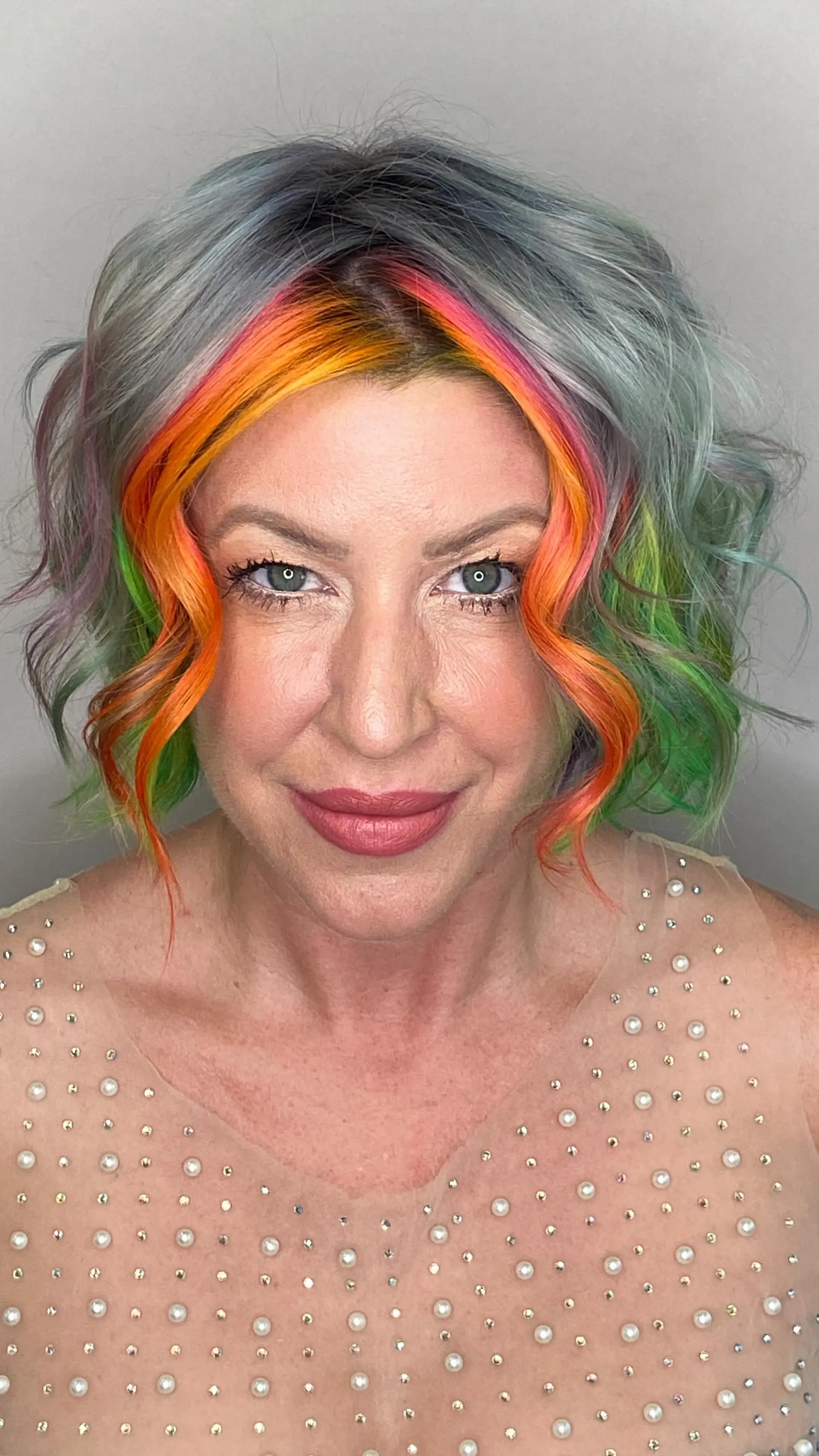 A textured chin-length wavy bob featuring a soft silver-grey base highlighted by vibrant sunset face-framing tones of neon pink and bright orange, with touches of lime green throughout. 1