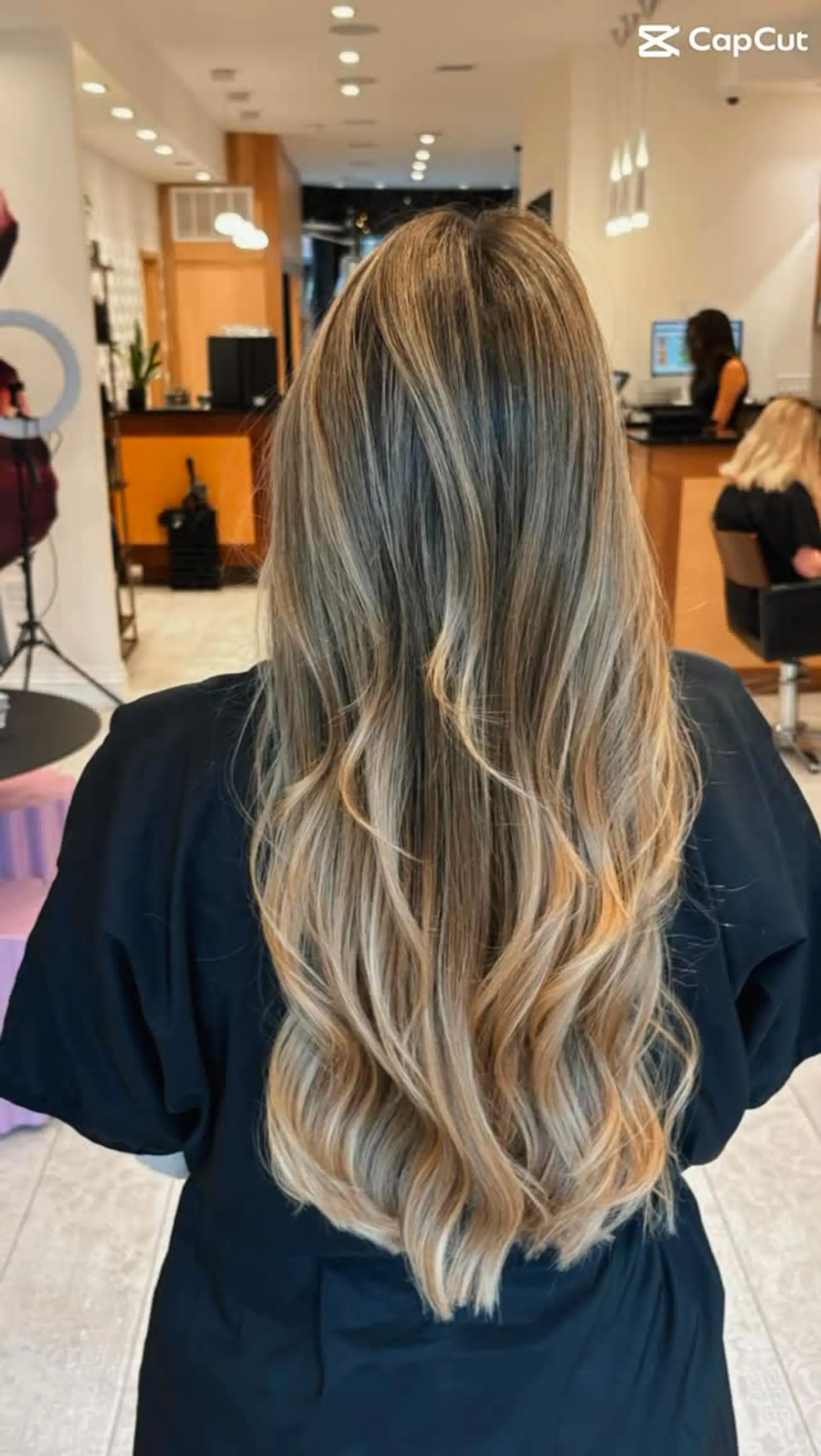 Long layered hair styled in soft, loose waves featuring a seamless dimensional blonde balayage over a darker root base. 1