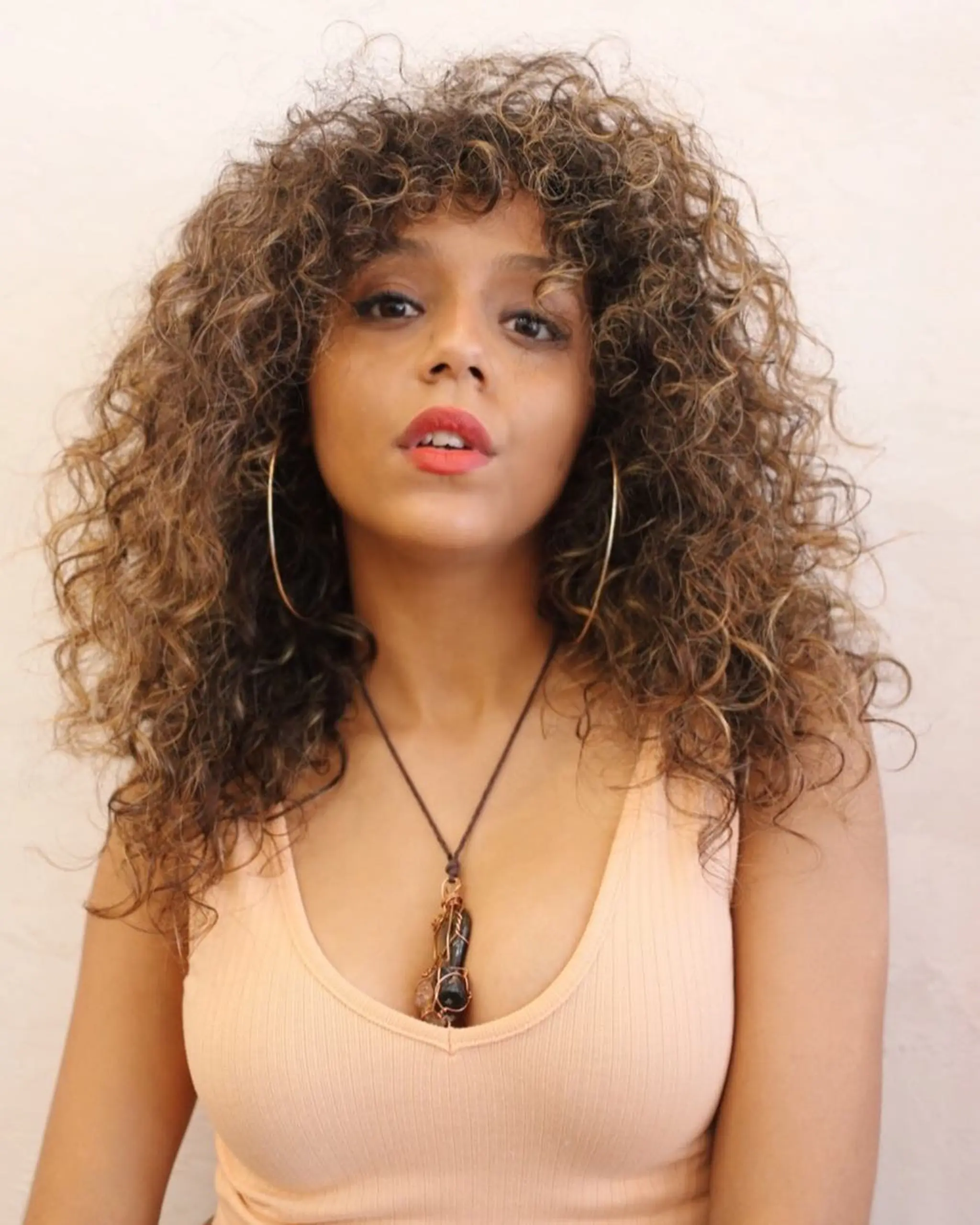 A vibrant and voluminous curly hairstyle with long, honey brown tresses interspersed with golden highlights. The curls are tight and full, cascading down past the shoulders. Curly bangs frame the face, adding a youthful vibe.