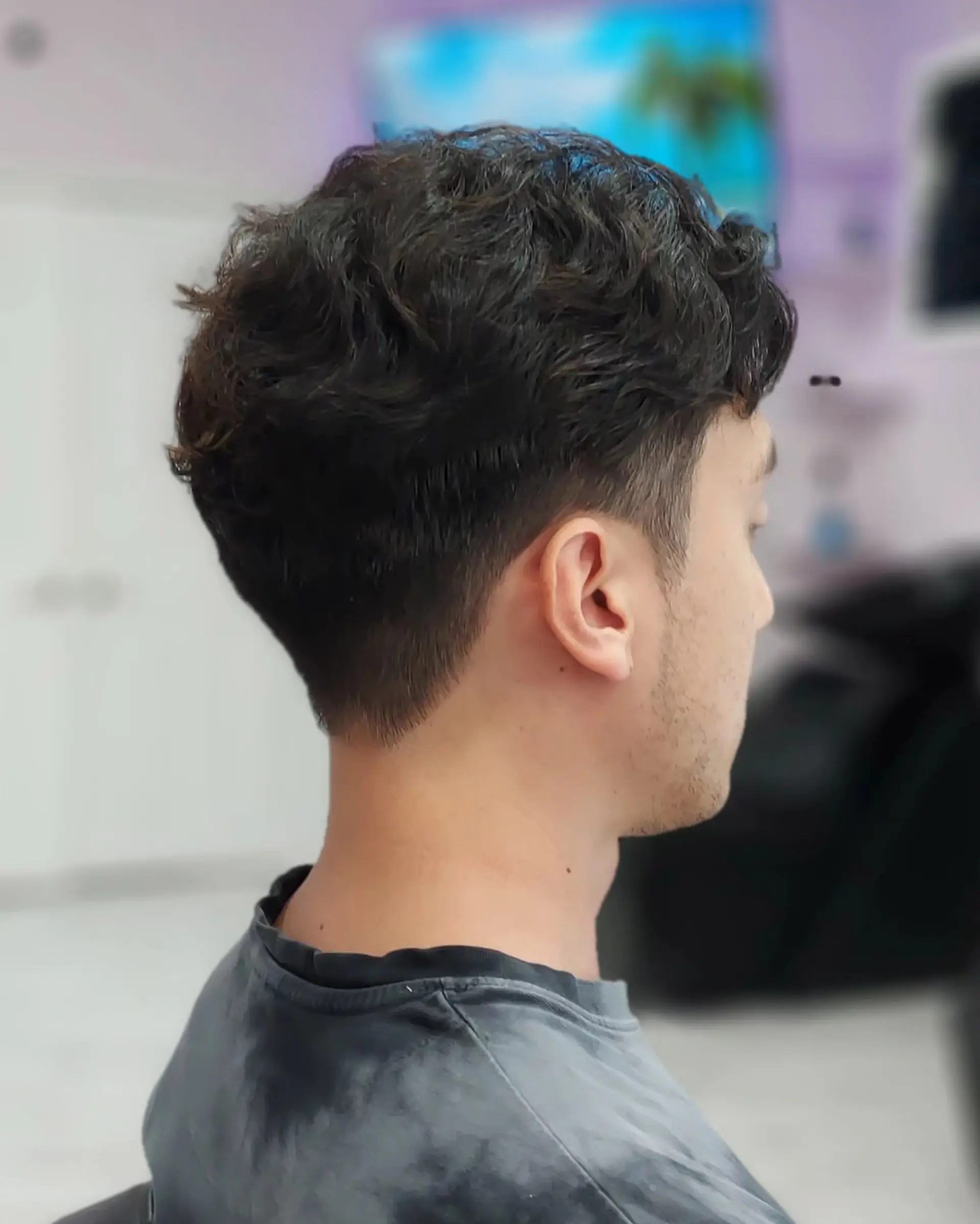 A men's short wavy perm styled with soft texture on top and a clean, subtle low taper around the neck and ears. 1