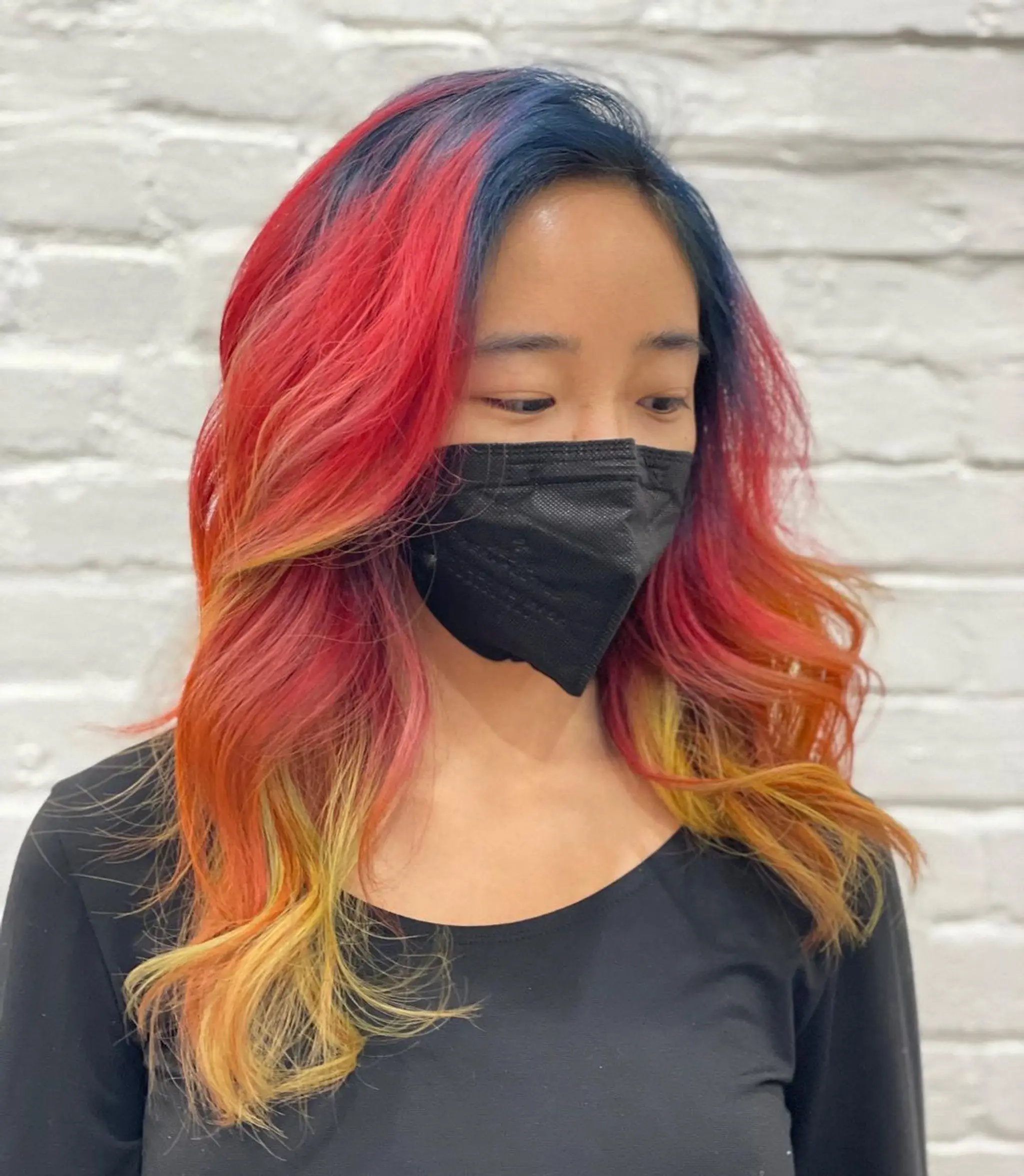 Long, wavy hair with a striking ombre gradient of red, yellow, orange, pink, and blue colors. The waves are loose and flowy, enhancing the layered haircut.