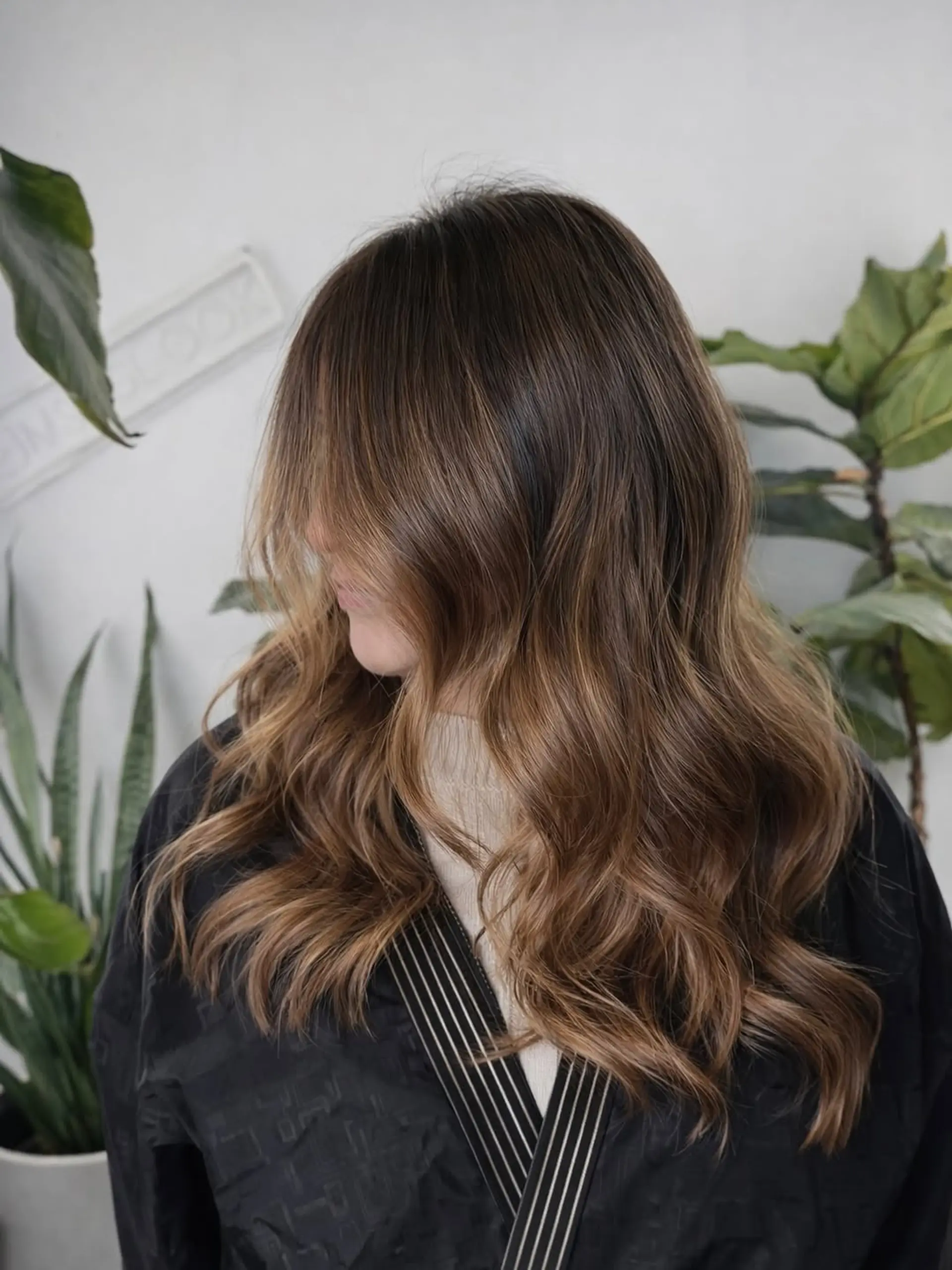 Long hair styled in soft, loose waves with a seamless balayage color technique, featuring lighter caramel tones blended into a dark brown base. 1