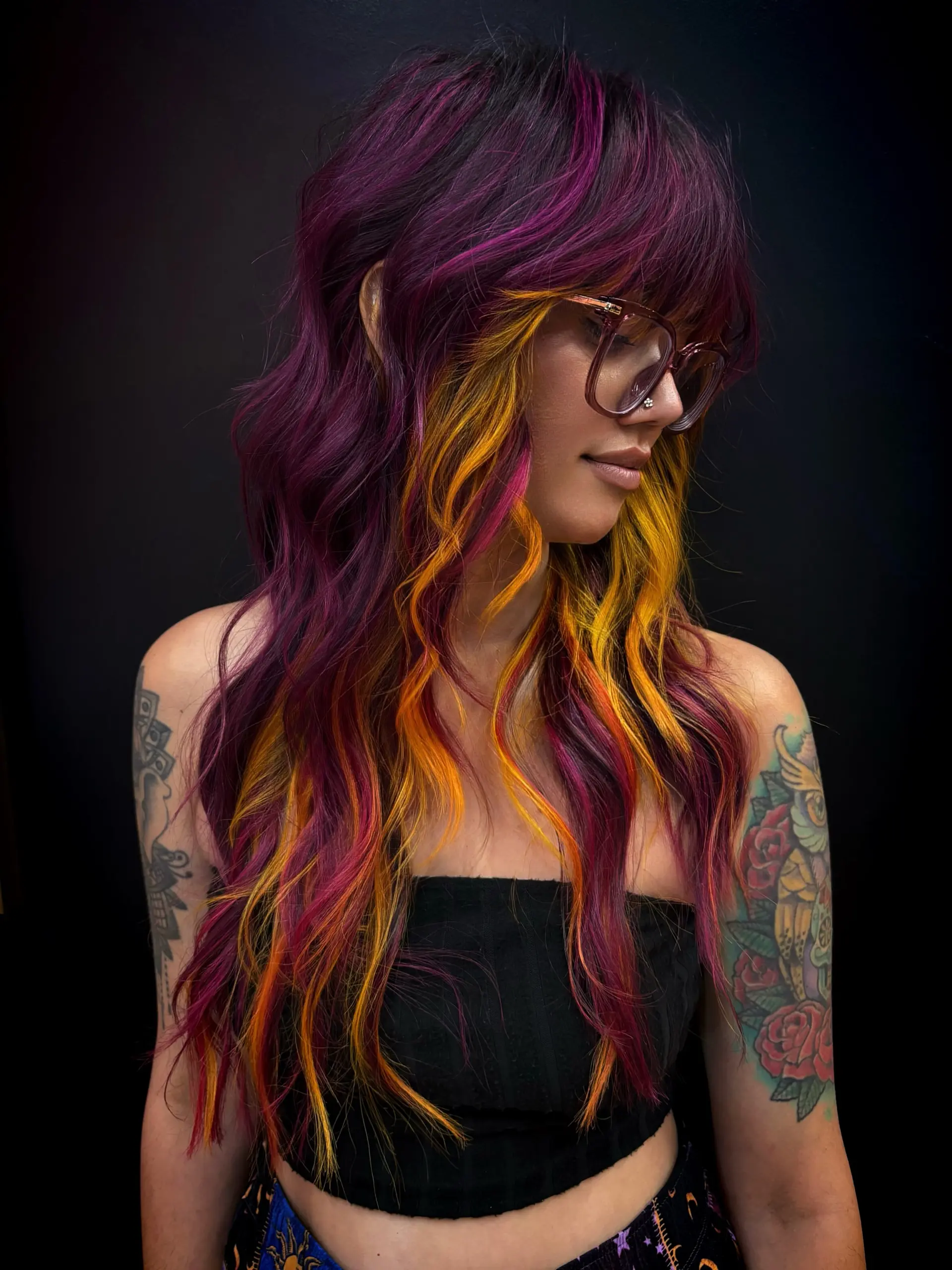A textured long modern shag with full fringe bangs, styled in loose waves with bold magenta-purple layers over warm yellow and orange underlights. 1