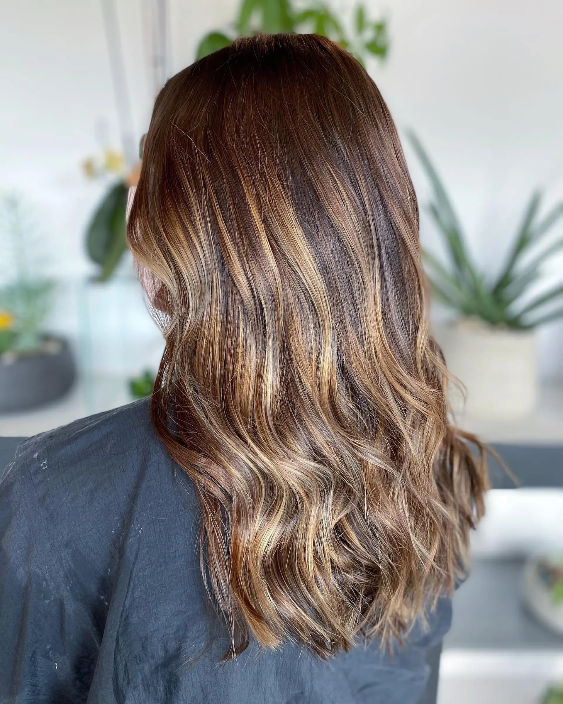Long dimensional brunette hair enhanced with warm caramel balayage highlights and styled into loose waves. 1