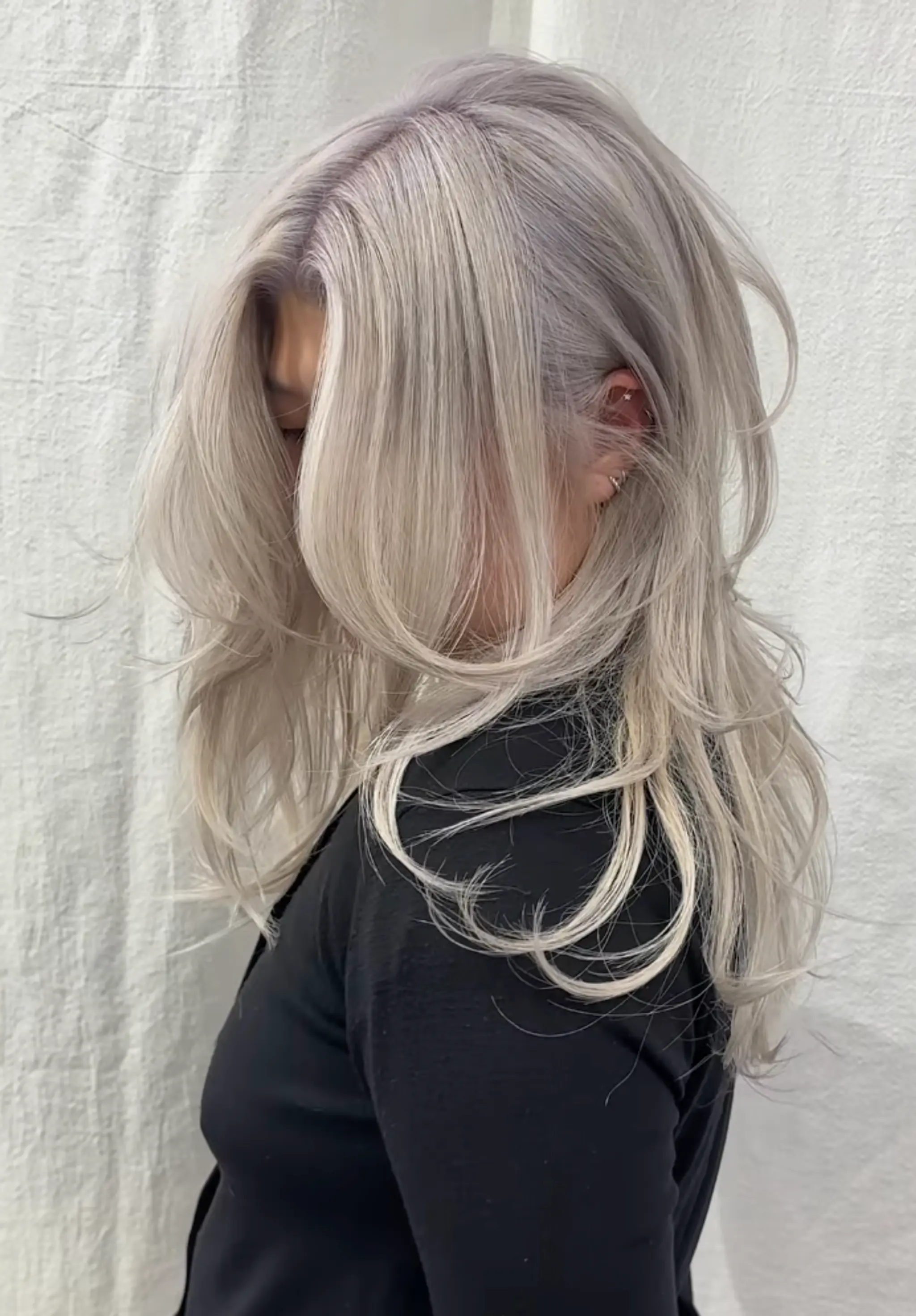 A long, wavy hairstyle featuring voluminous layers and a side part. The platinum blonde and silver hues create a cool-toned look, with face-framing waves and textured ends for added dimension.