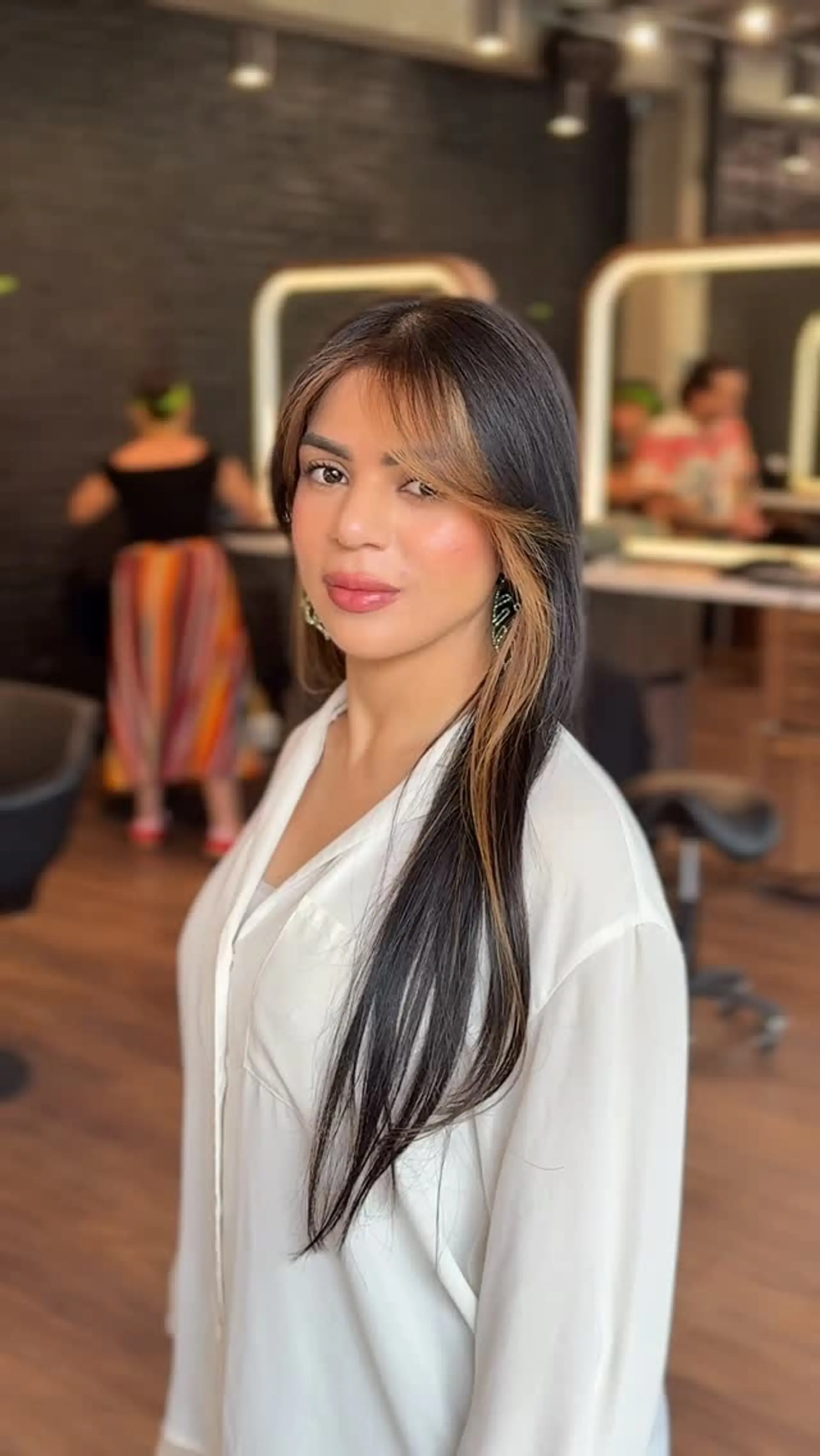 Long straight dark brown hair styled with curtain bangs and a warm honey blonde face-framing money piece. 1