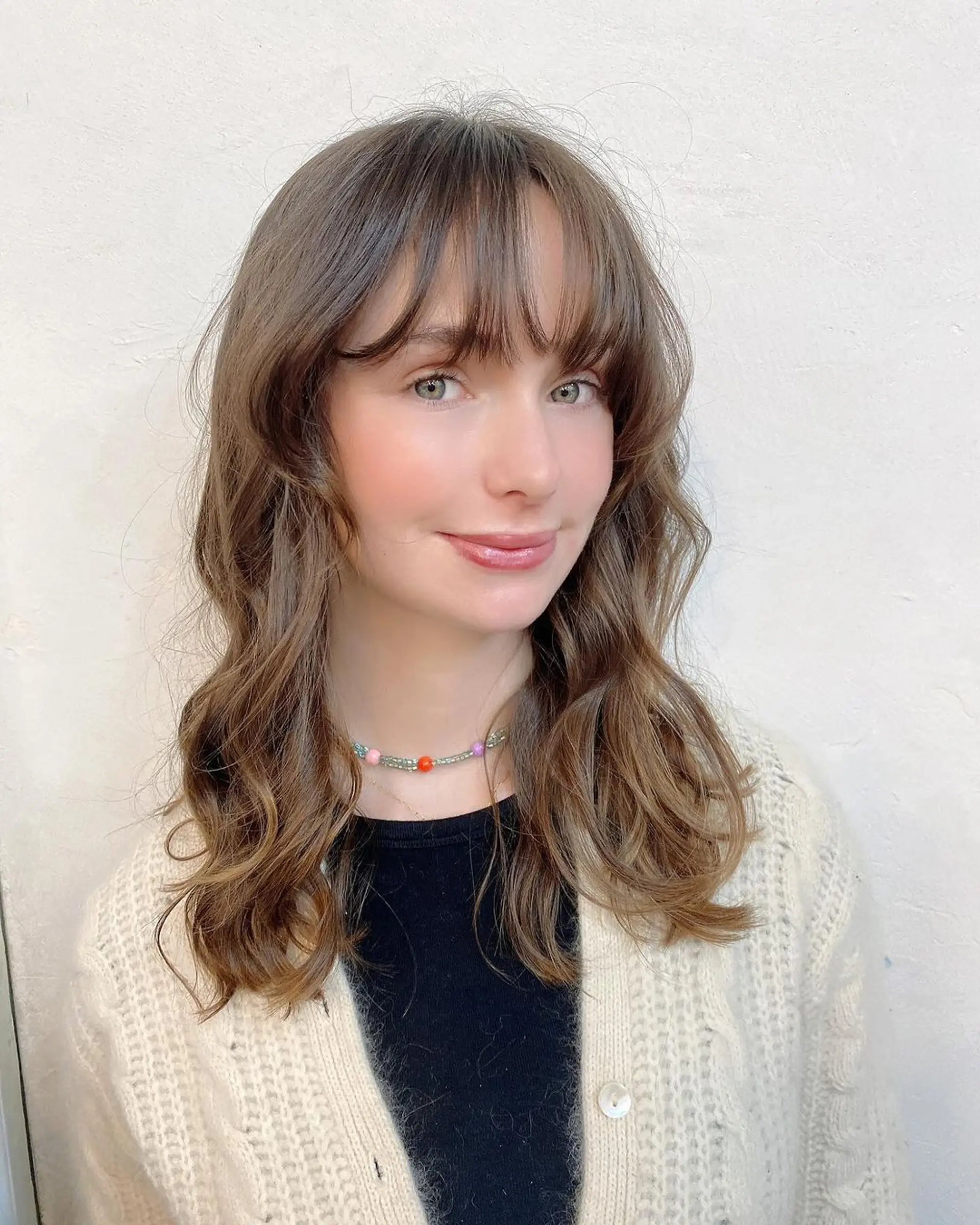 A soft, medium-length wavy hairstyle with light brown hair cascading in gentle waves. Wispy bangs graze the eyebrows, adding to the soft look. Layering throughout enhances movement and volume, maintaining an elegantly tousled appearance by Coco. 1