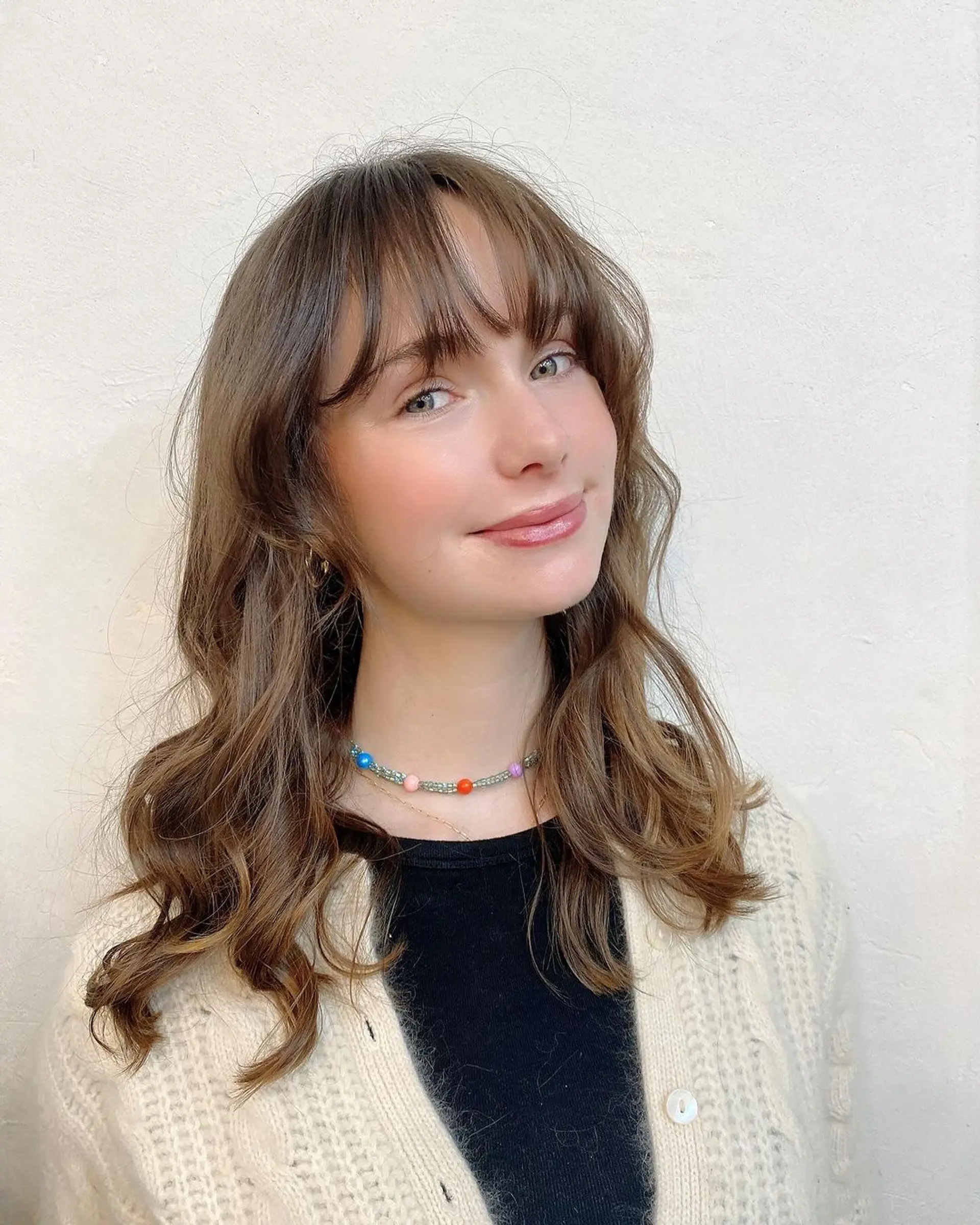 A soft, medium-length wavy hairstyle with light brown hair cascading in gentle waves. Wispy bangs graze the eyebrows, adding to the soft look. Layering throughout enhances movement and volume, maintaining an elegantly tousled appearance by Coco. 2