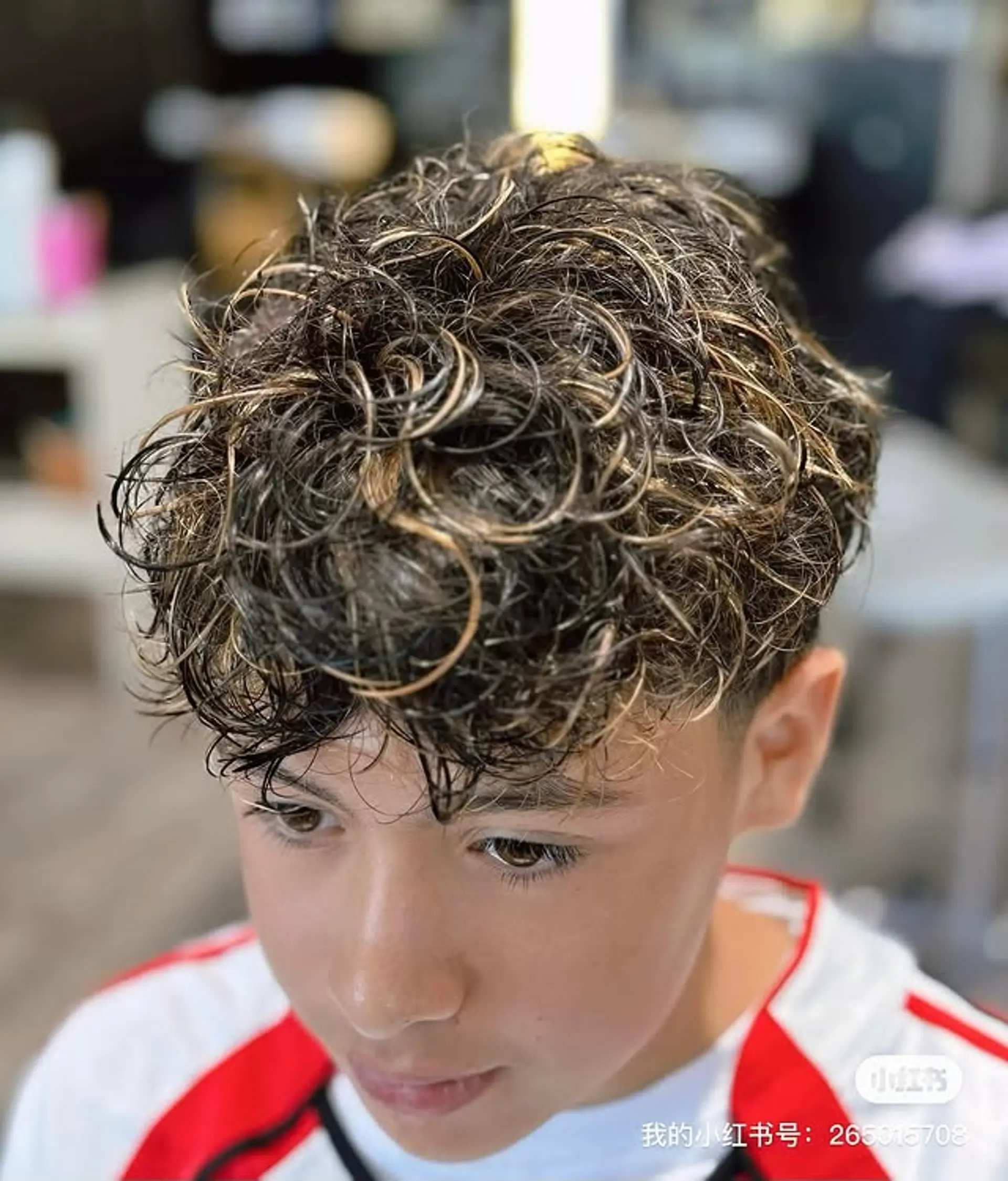 A modern men's cut with a lively, textured curly top that boasts prominent blonde and caramel highlights, contrasted by shorter hair on the sides. This style emphasizes natural curl patterns and adds dimension with strategic coloring. 1