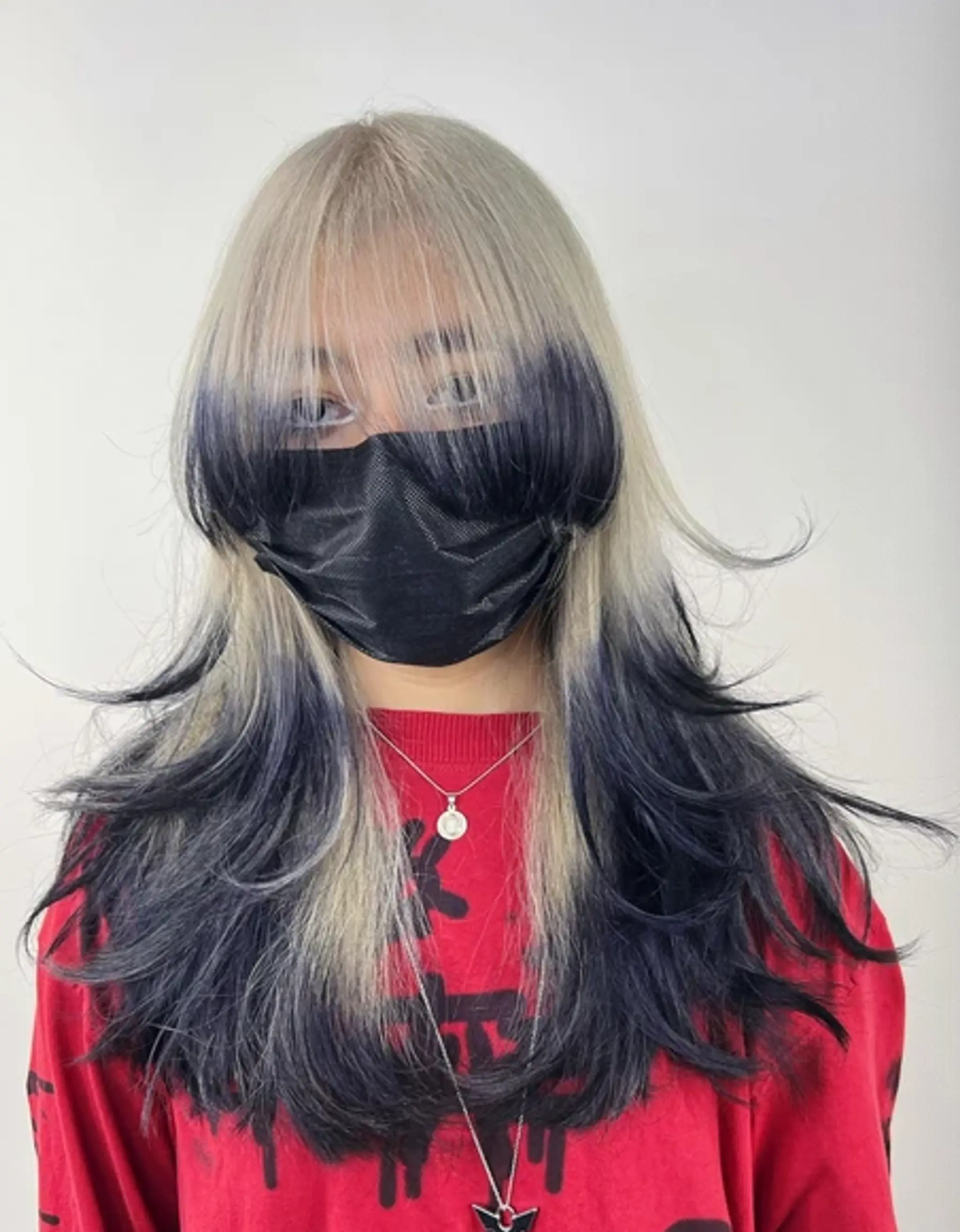 A bold two-tone ombre hairstyle with platinum blonde roots fading into deep black, complementing long, layered wavy hair. Features wispy bangs that frame the face for a modern and edgy look.