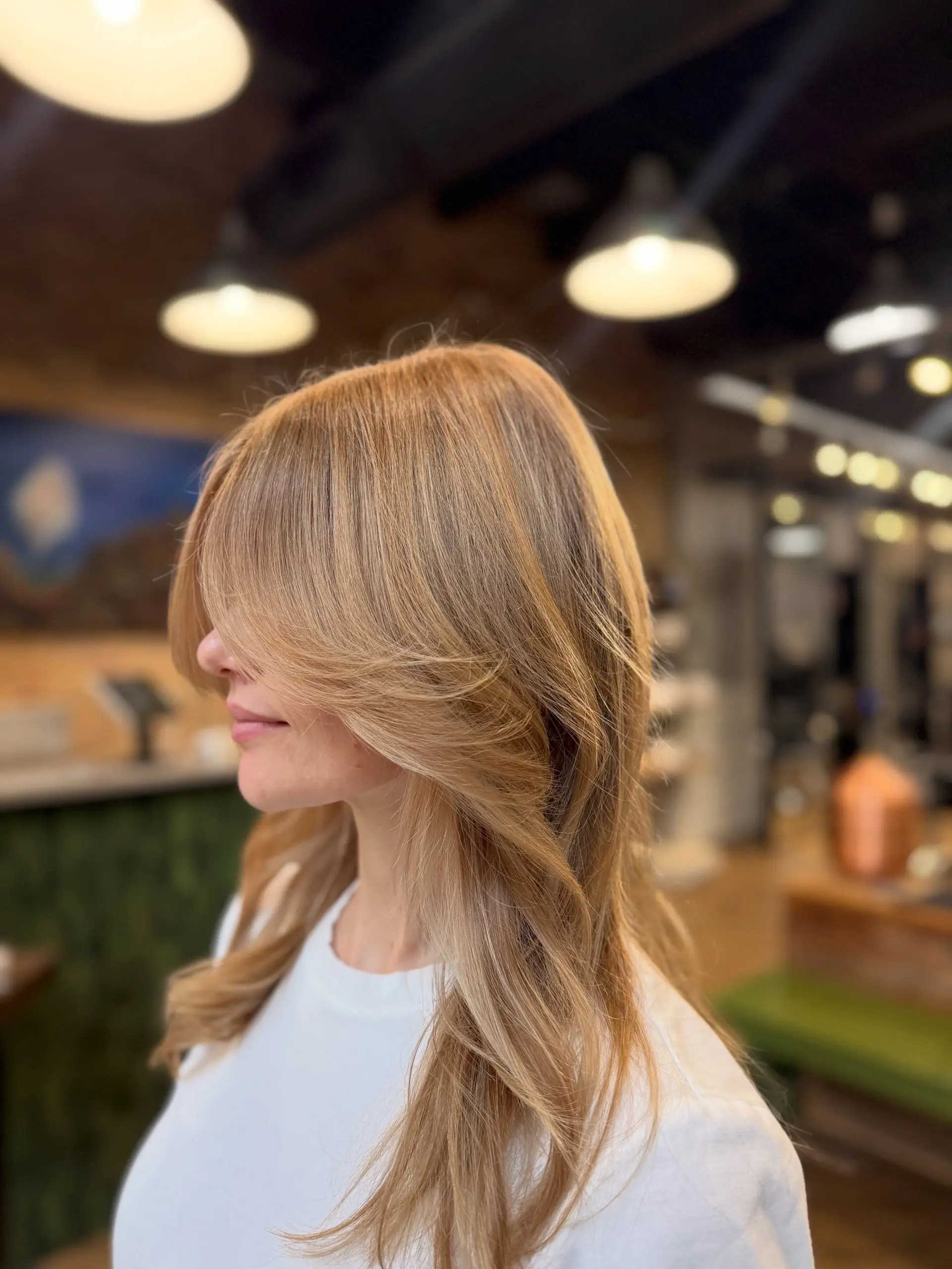 A long warm honey blonde haircut featuring long, face-framing curtain bangs styled with soft, voluminous movement. 1