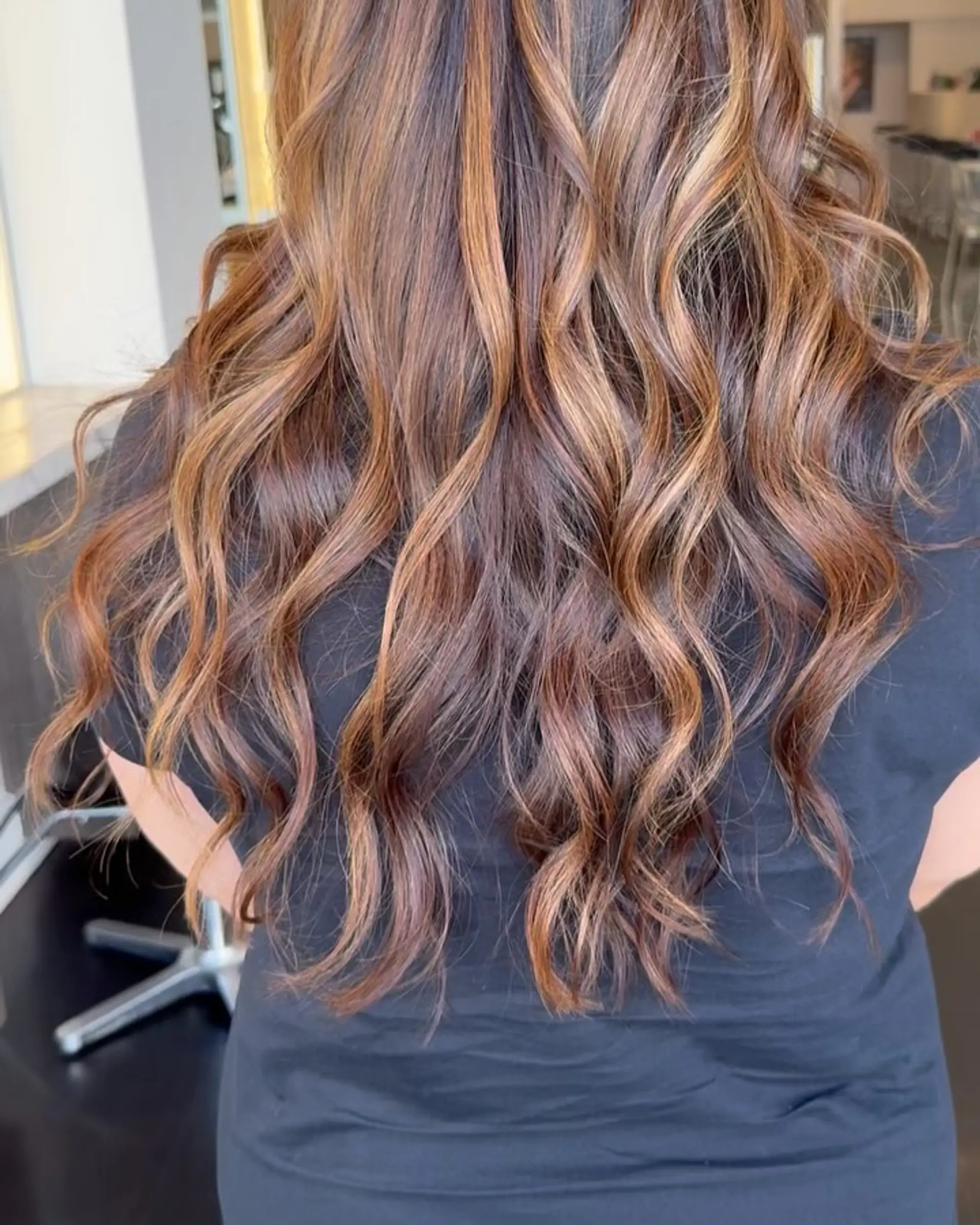 Long dark brown hair featuring multi-dimensional caramel and warm honey highlights, styled into soft textured waves. 1