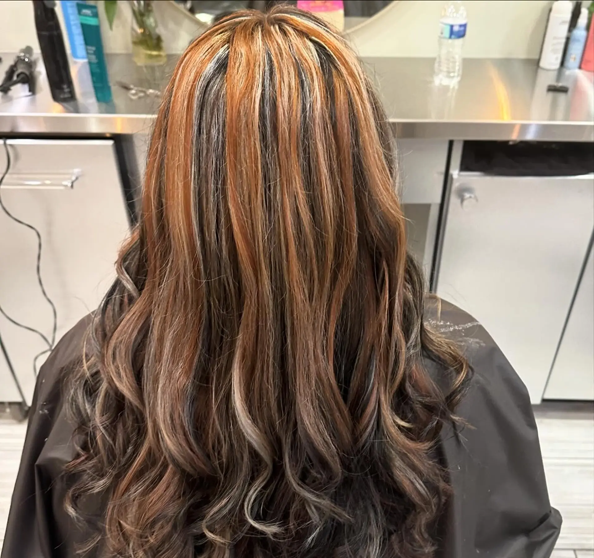 Long wavy hair styled with bold, high-contrast chunky highlights in copper and warm blonde over a dark brunette base. 1