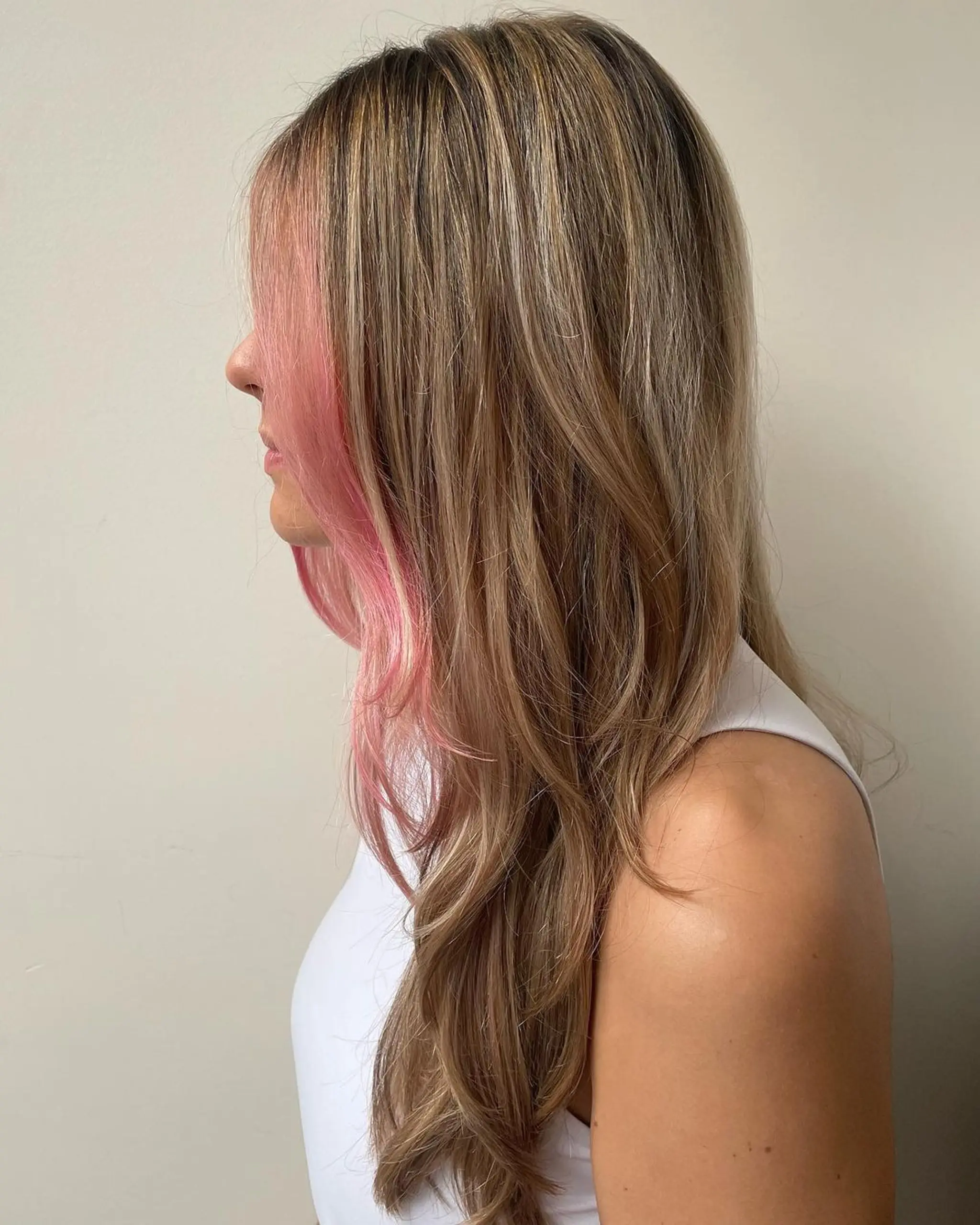 A long, layered hairstyle with a natural blonde base and a pink streak that frames the face, featuring soft, flowing waves.