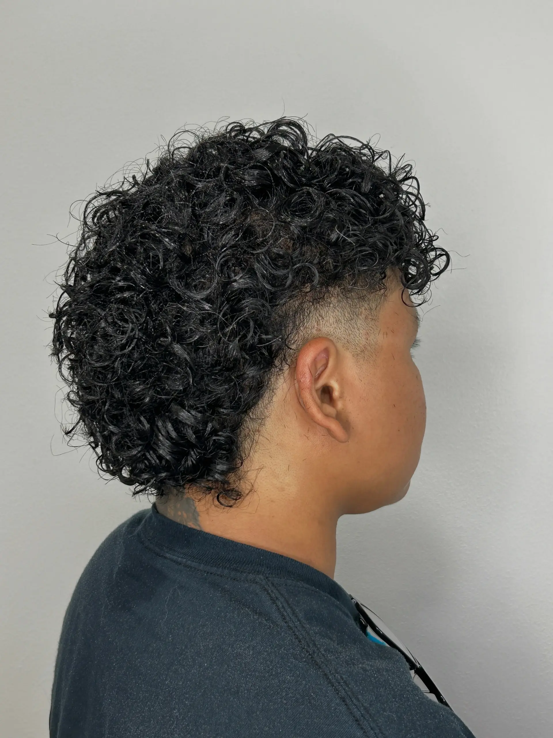 A modern curly mullet featuring defined, wet-look curls on top and down the neck paired with a clean taper fade around the ear. 1
