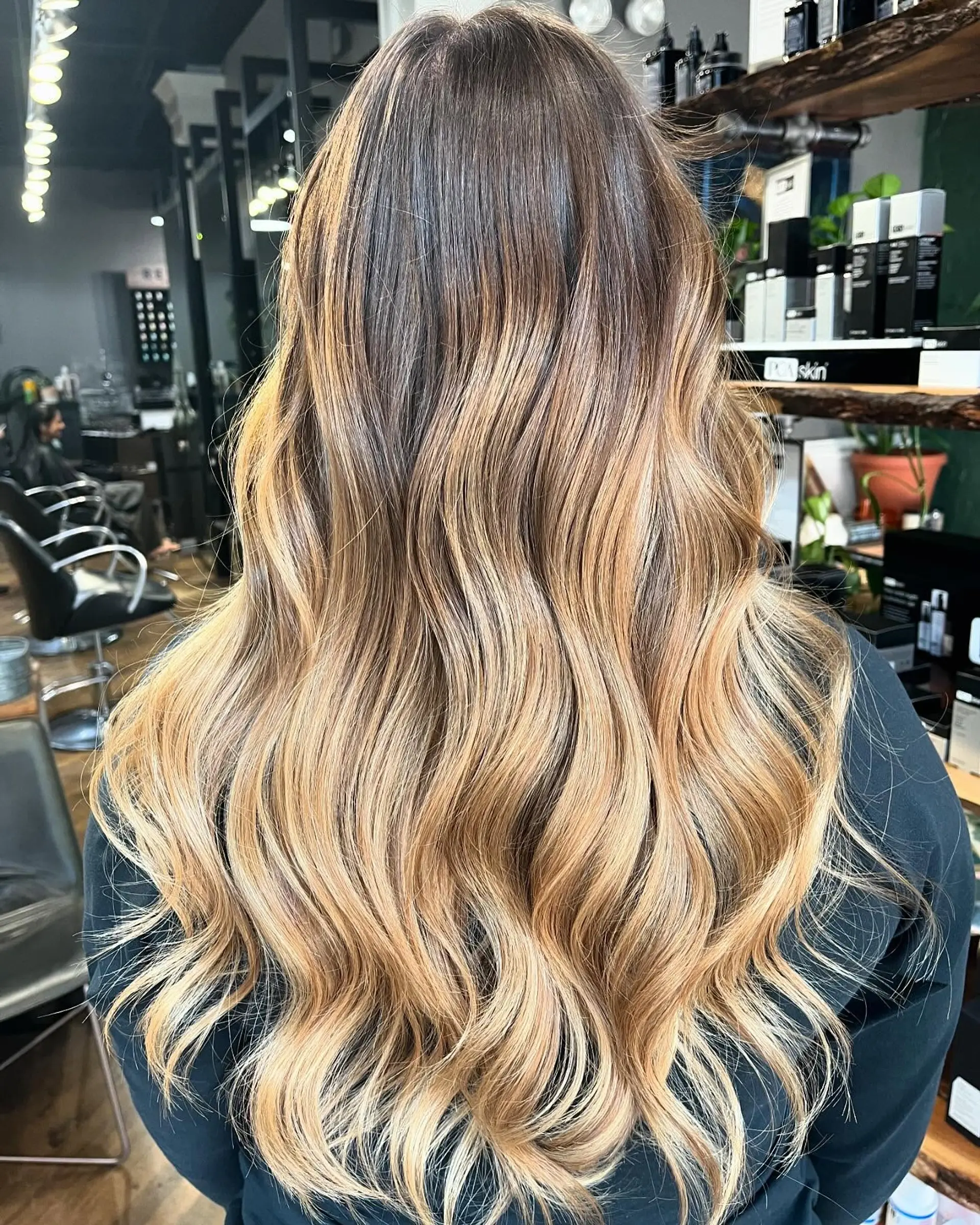 Long dimensional balayage transitioning from deep brunette roots into warm honey and golden blonde ends styled in soft waves. 1