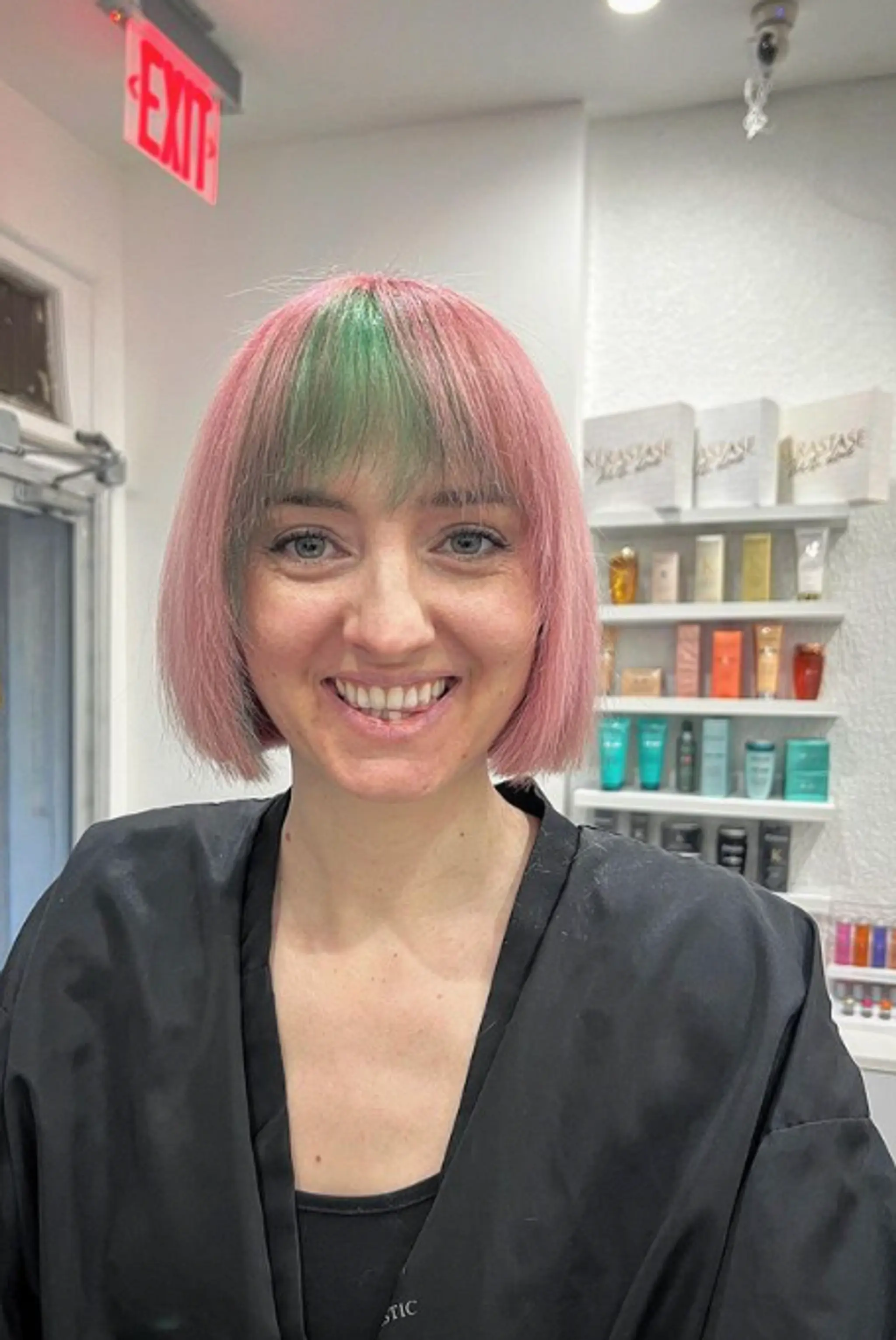 A straight, blunt-cut bob that falls just below the chin, featuring straight, full bangs across the forehead. The primary color is a soft pastel pink, complemented by a green accent in the bangs.