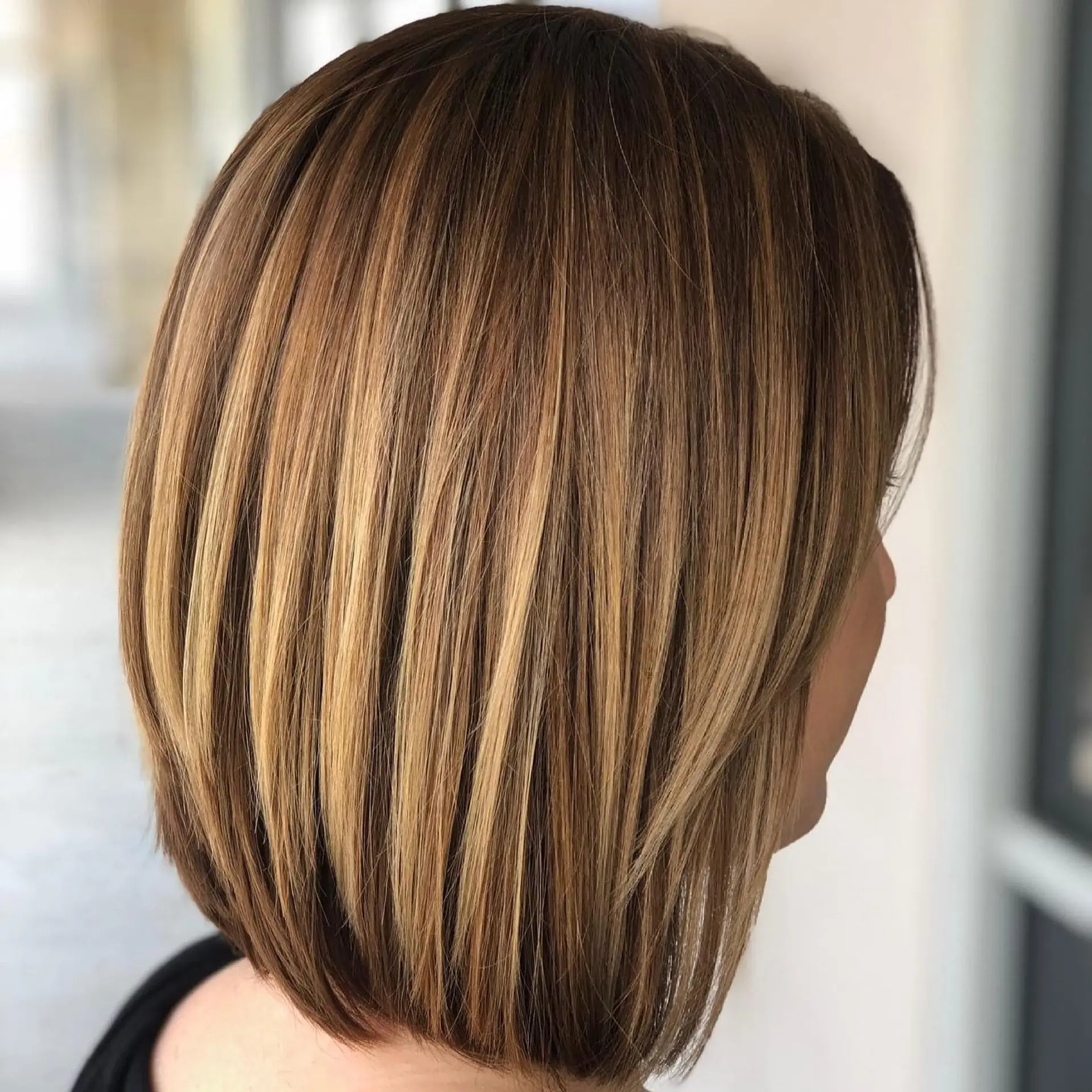 A stylish A-line bob with subtle layering, longer in the front and gradually shorter towards the back, enhanced with dimensional caramel and golden blonde highlights over a dark brown base. 1