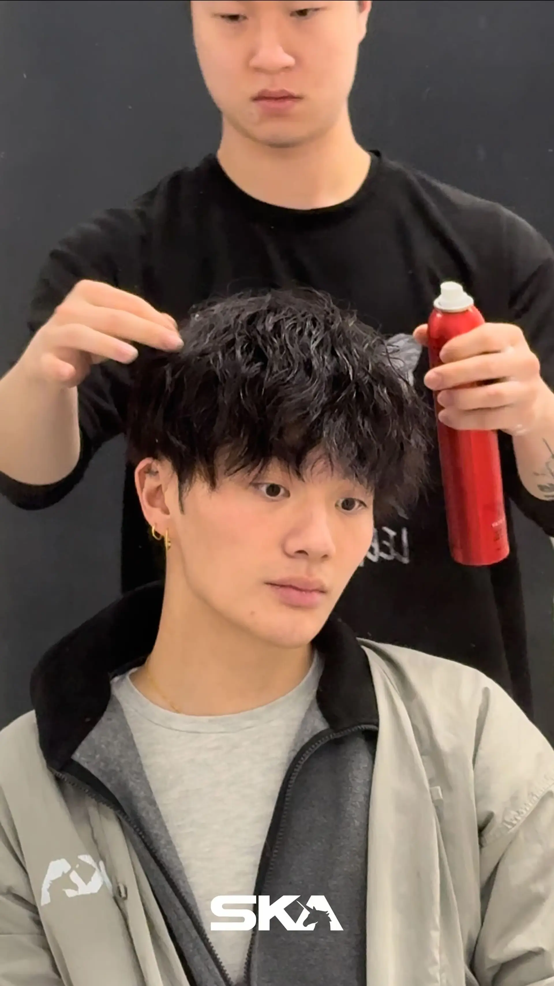 A men's short layered haircut enhanced with a textured perm, styled forward with defined, piecey waves and full fringe. 1