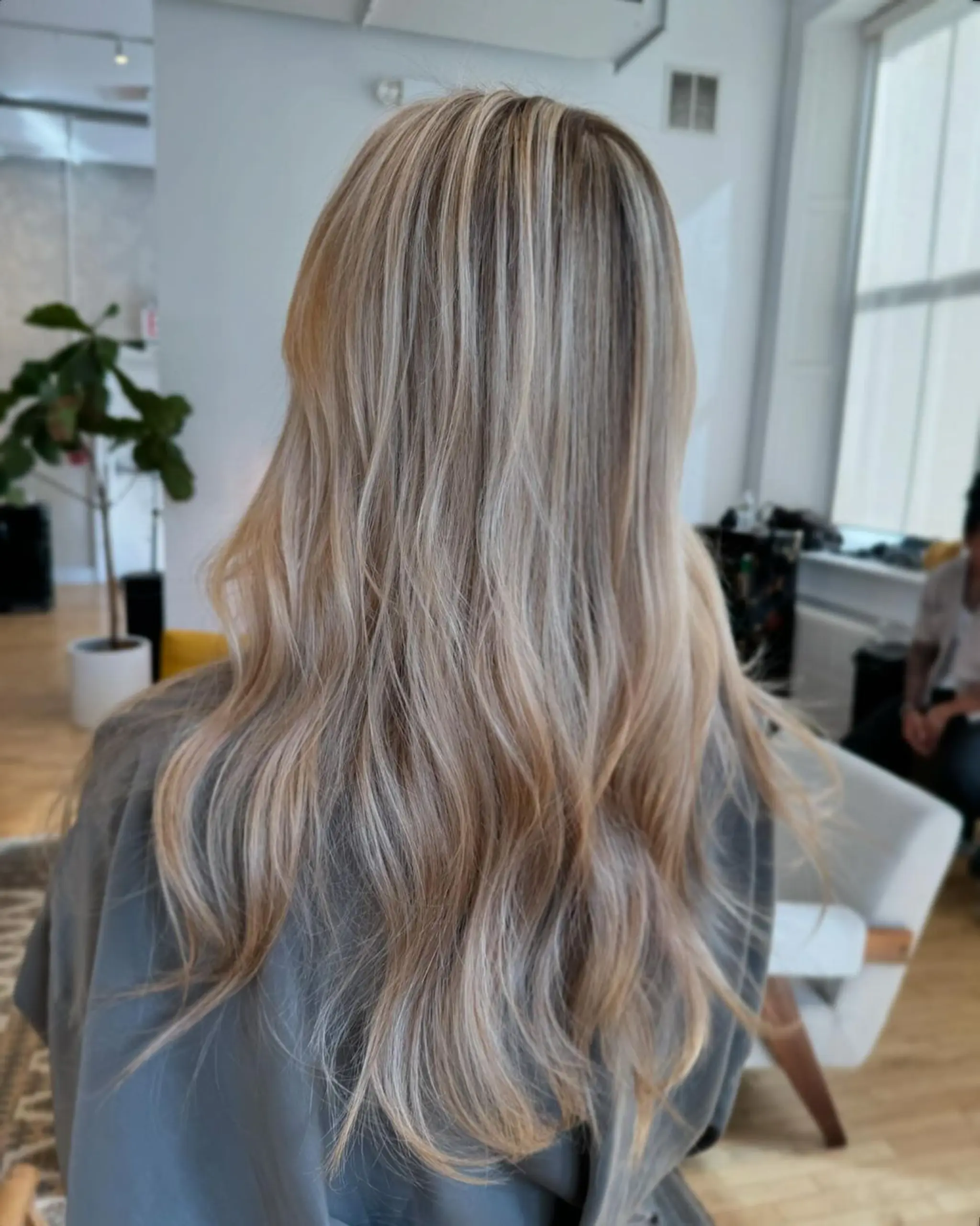 Long, wavy hair with a blend of blonde, platinum, and honey hues. Soft, natural waves with sun-kissed highlights and multi-tonal colors. Layered cut for enhanced flow and movement.