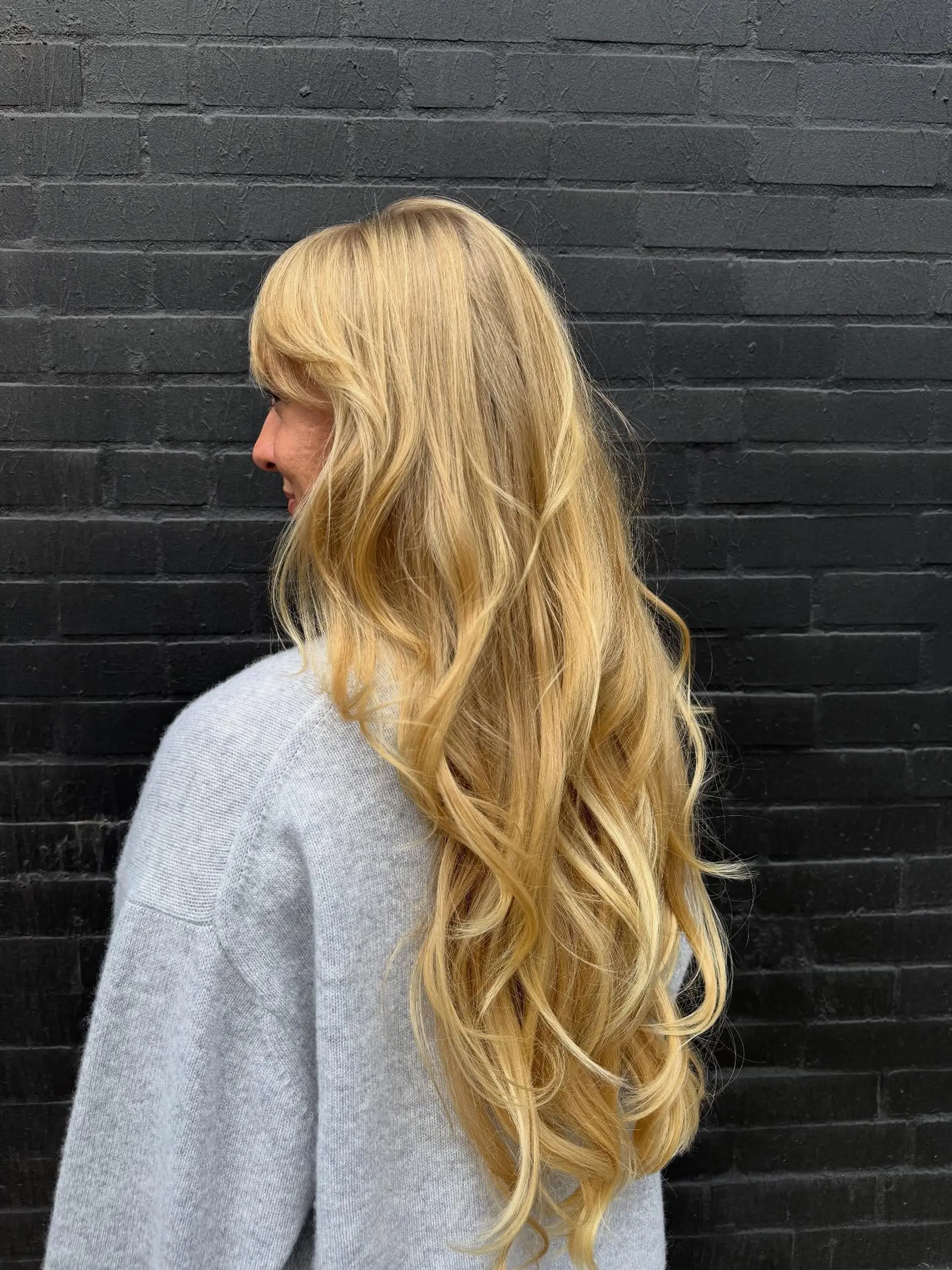 Very long, warm golden blonde hair cut with soft cascading layers and styled into loose waves with side-swept bangs. 1