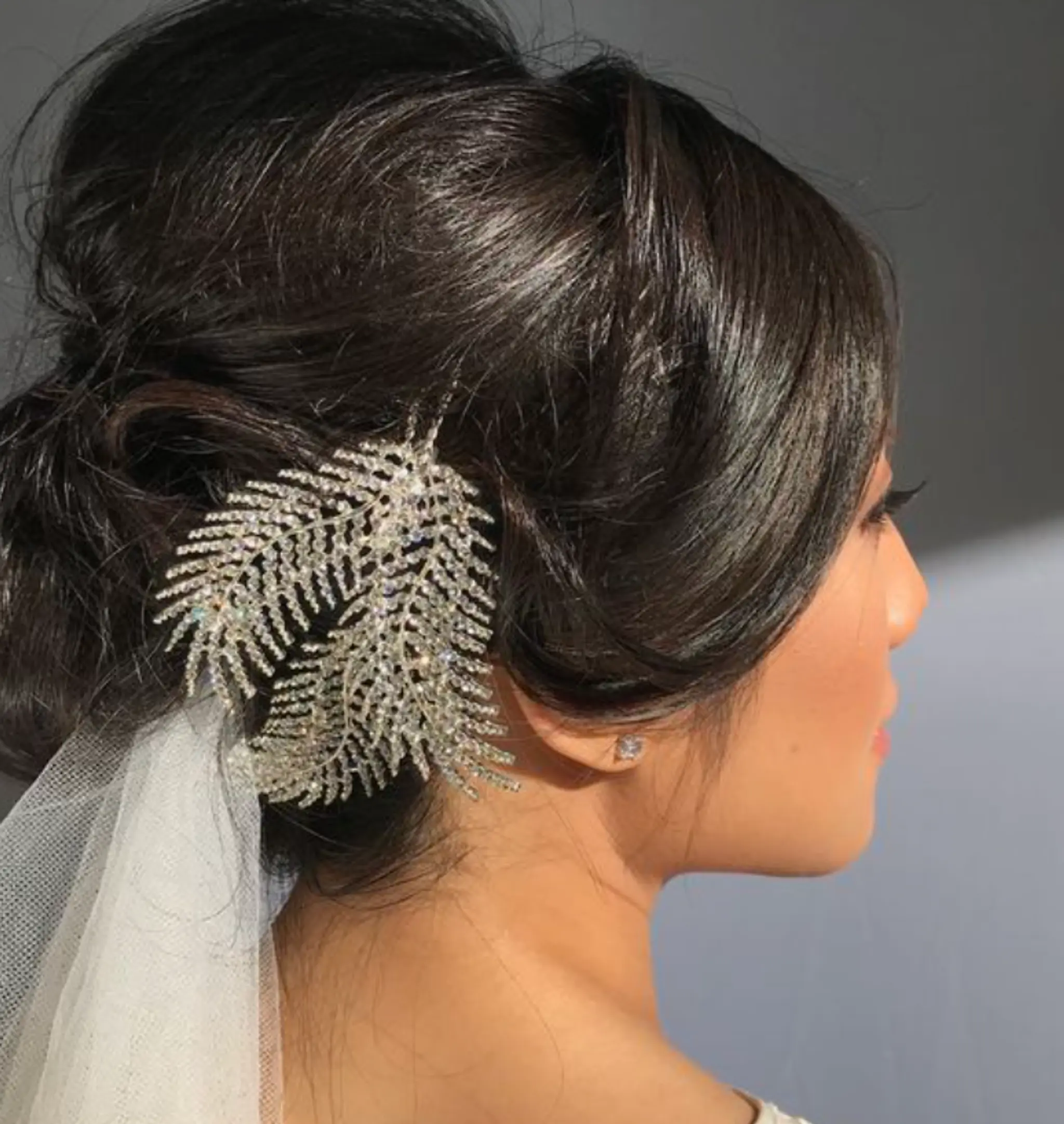 A sophisticated updo hairstyle with long, straight black hair gathered into a sleek bun, adorned with a sparkling leaf hair accessory and a white bridal veil.