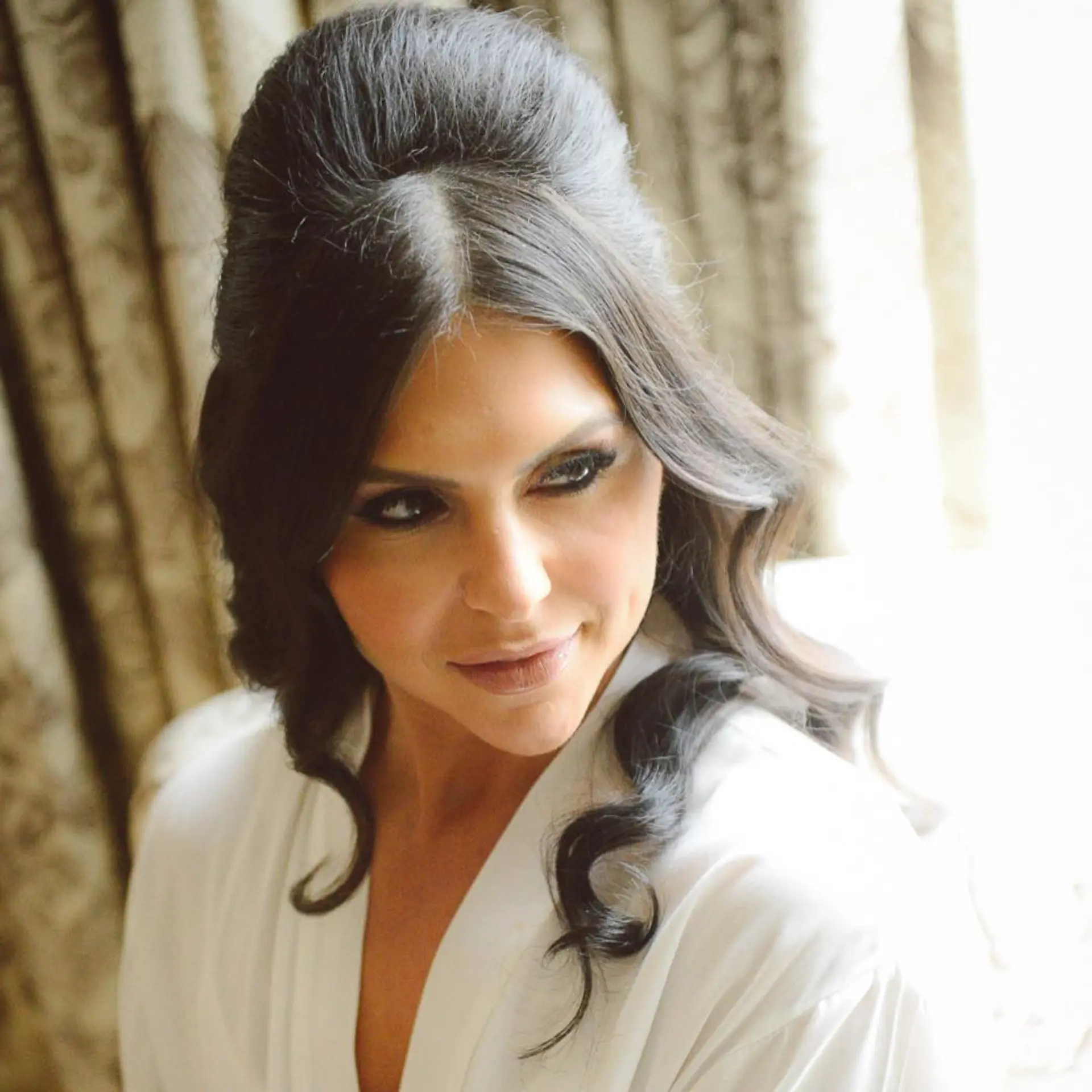 A dramatic retro-glam bridal half-updo featuring a voluminous teased bouffant crown with center-parted face-framing strands and soft spiraled curls falling over the shoulders. 1