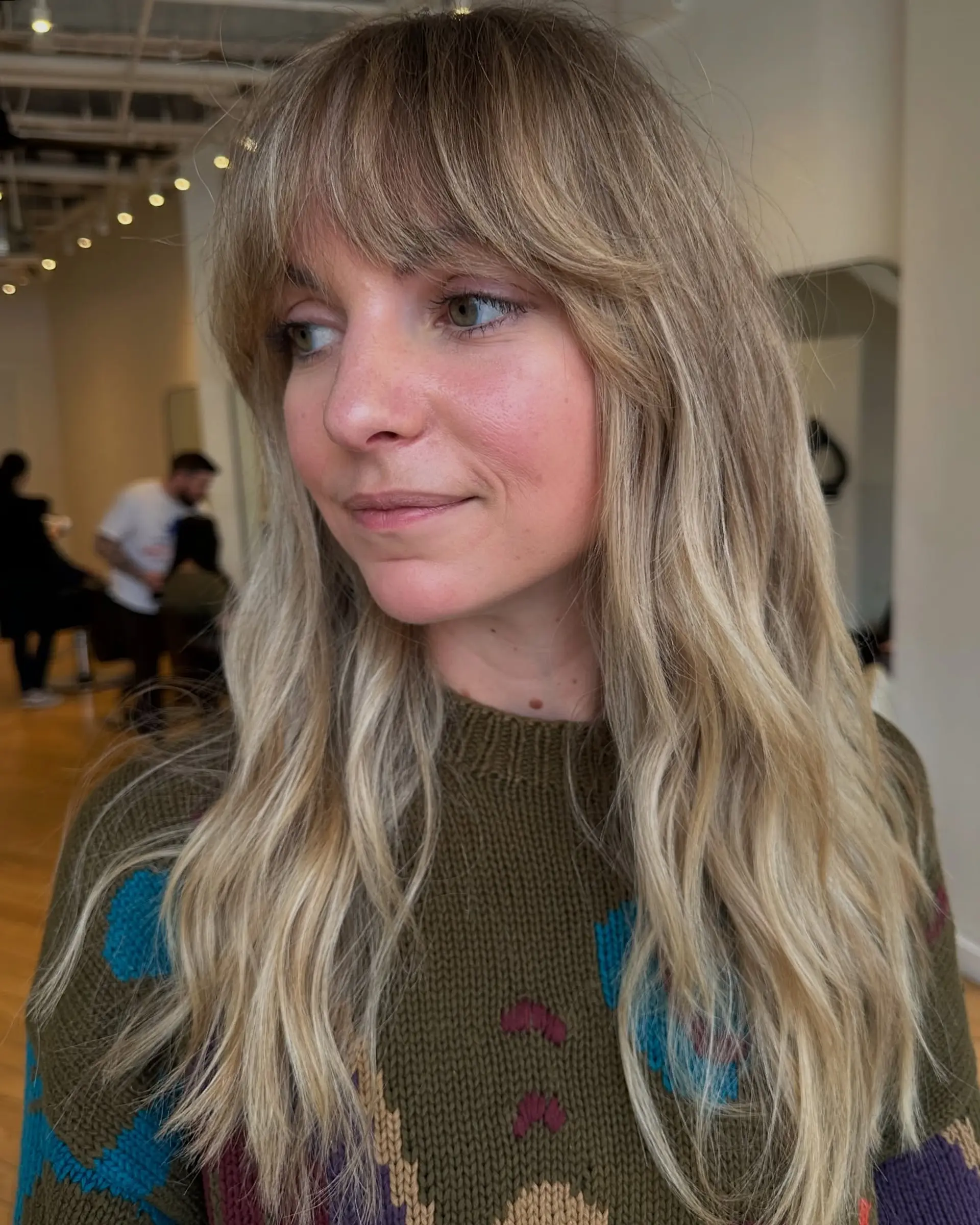Long wavy blonde hair cut with dimensional highlights and soft, eyebrow-skimming curtain bangs. 1