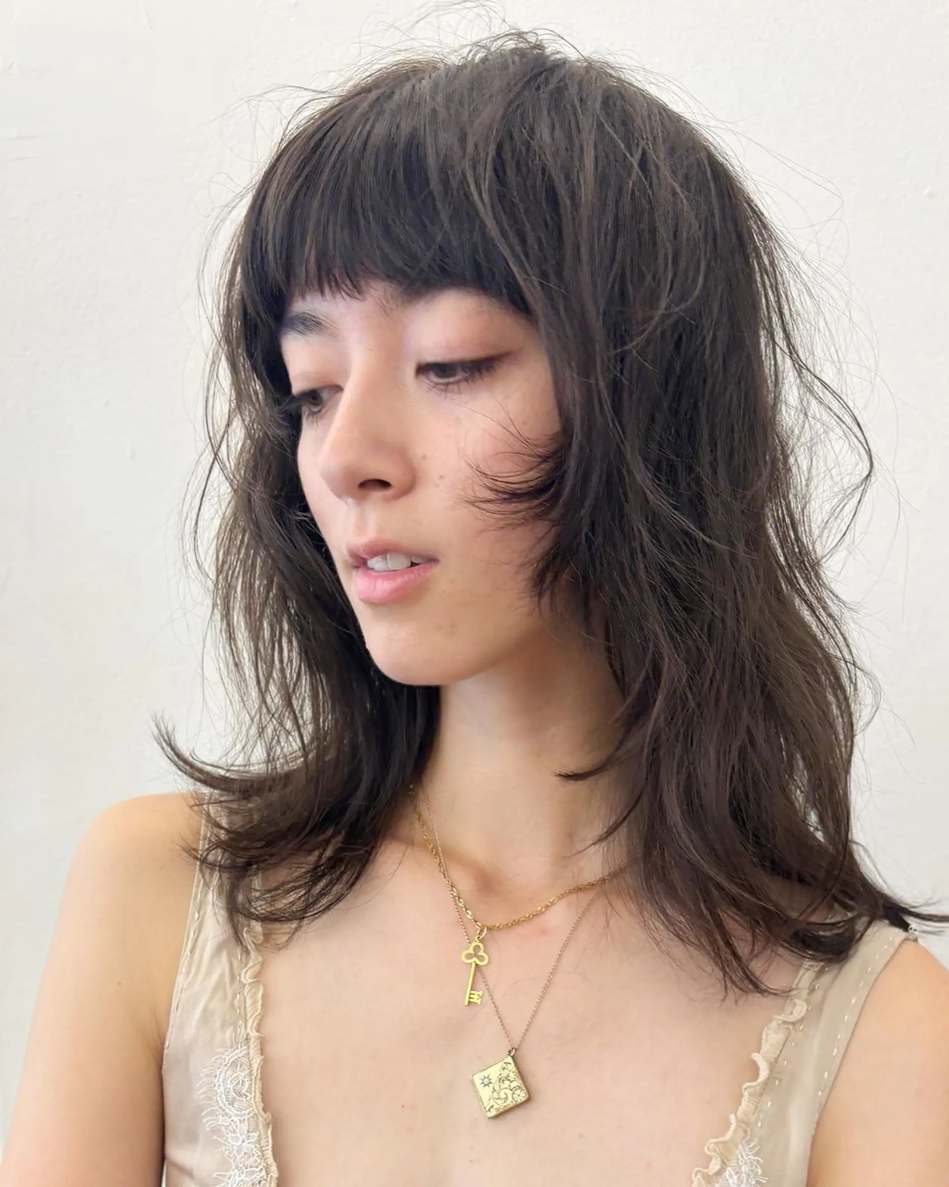 A medium-length, wavy dark brown hairstyle with textured, tousled layers, a shaggy cut, and blunt bangs. 1