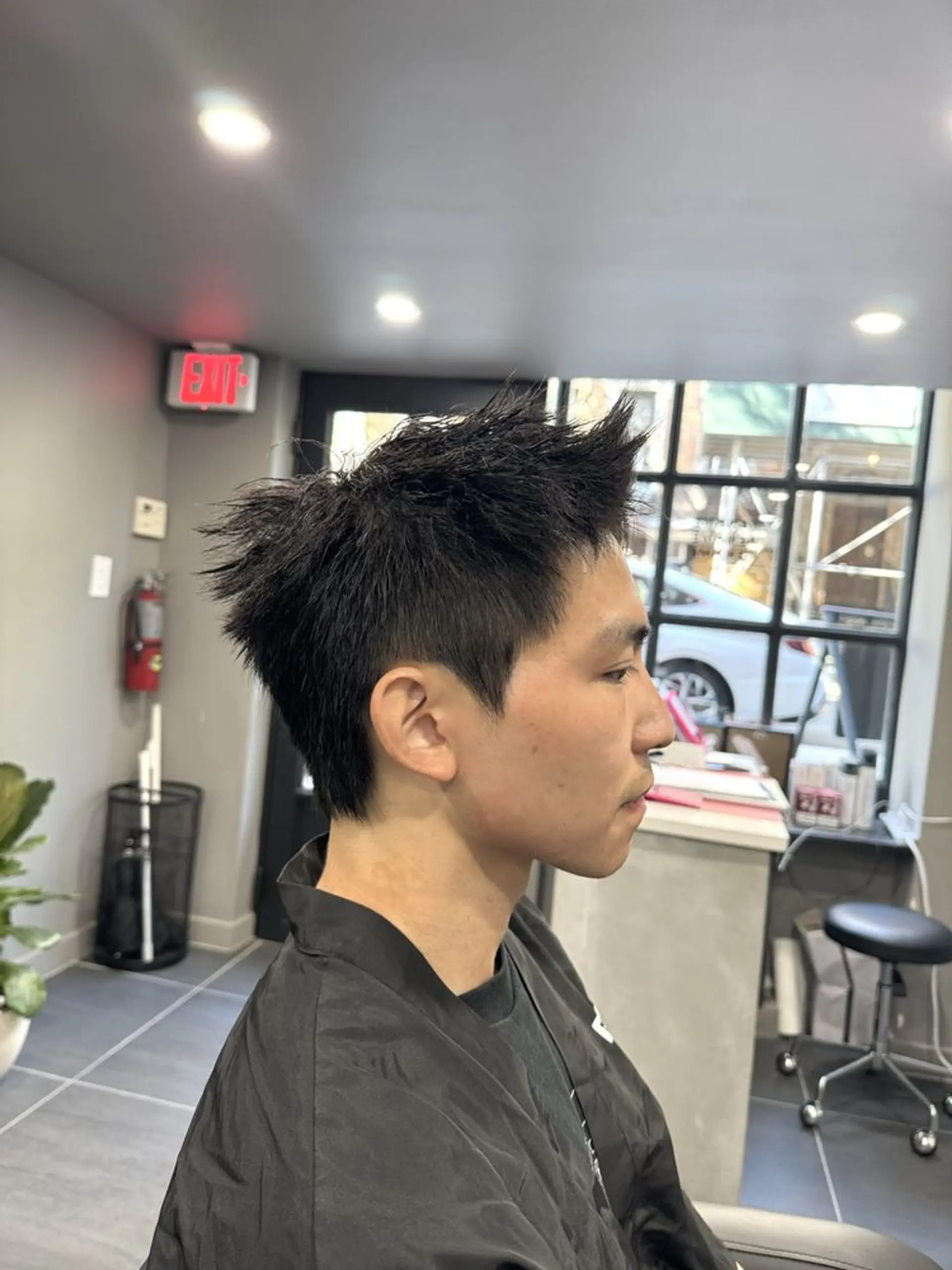 This hairstyle features a modern, edgy look with straight, short black hair styled into sharp spikes. The overall texture is choppy and filled with volume, giving it a dynamic and contemporary appearance. The sides are faded, adding to the sleek and clean aesthetic. Down perm used on the side and perm on the top to accentuate the texture of the cut. 1