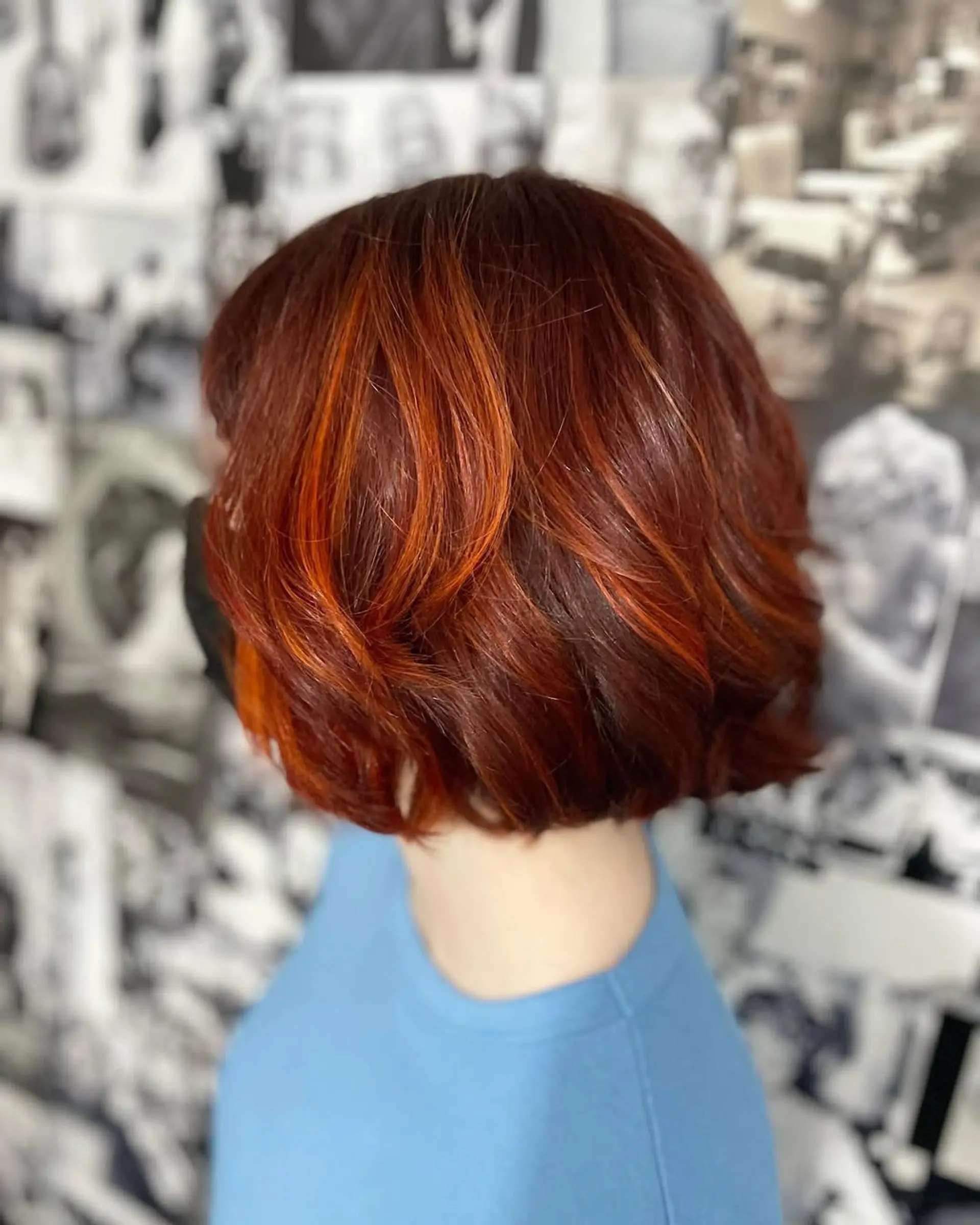 A short, textured bob hairstyle characterized by its rich auburn red base color, artfully enhanced with bright copper and orange dimensional highlights. The hair is styled with soft waves, providing volume and a contemporary finish. 1
