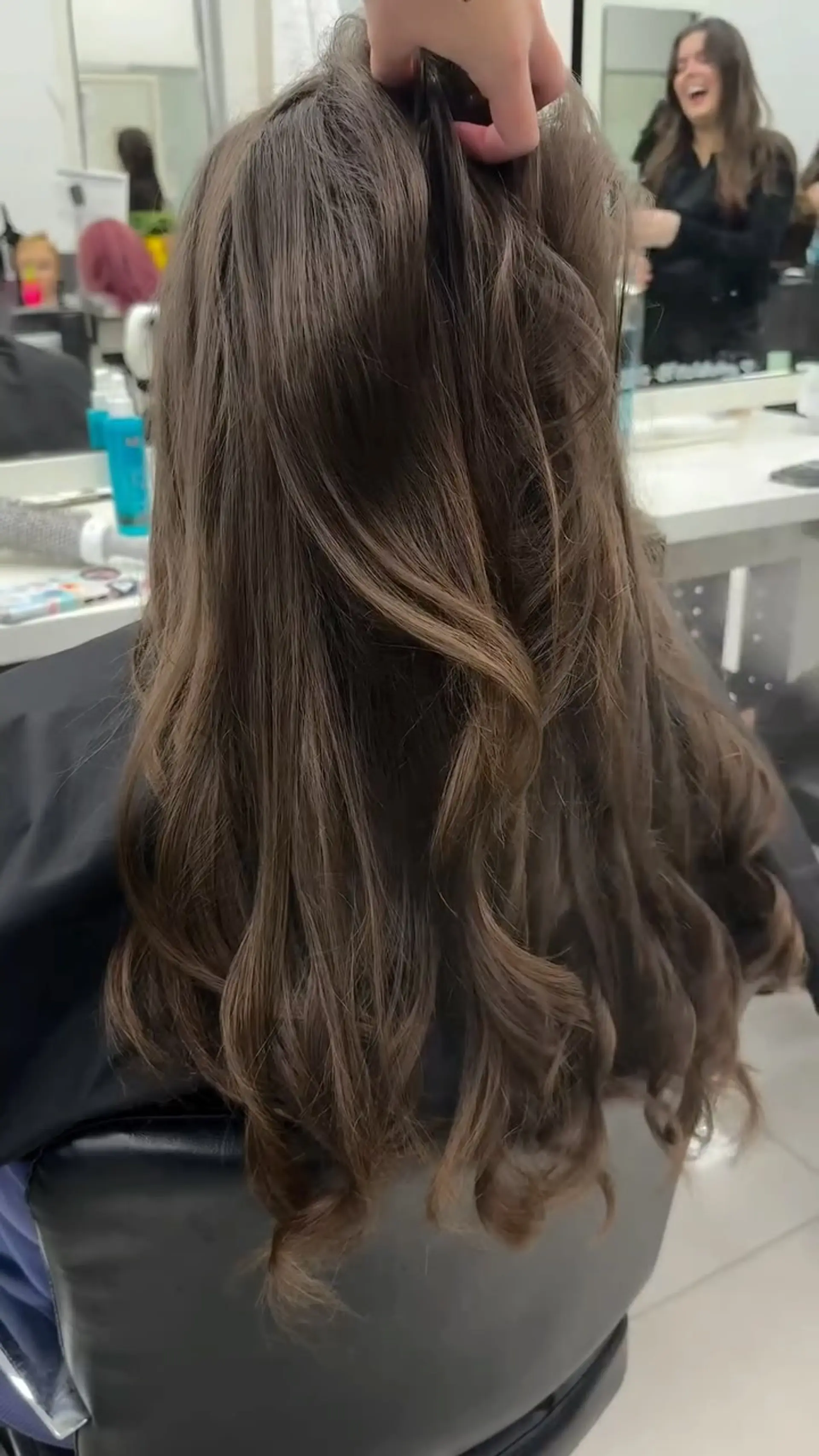 Long medium-brown hair styled with soft, cascading waves and polished curled ends. 1