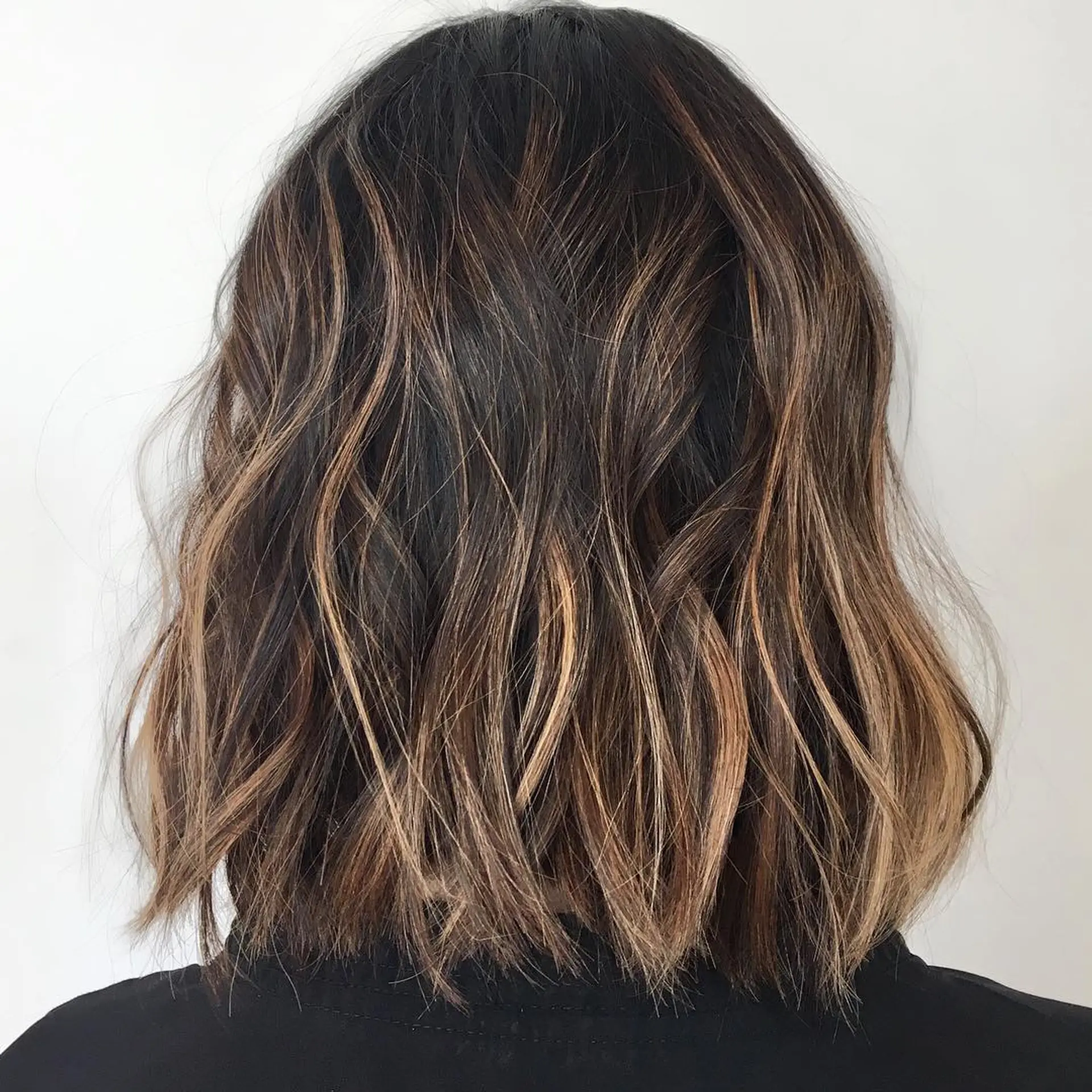 A blunt, textured medium-length lob featuring soft, tousled waves with seamless caramel foilayage highlights over a rich dark brown base. 1