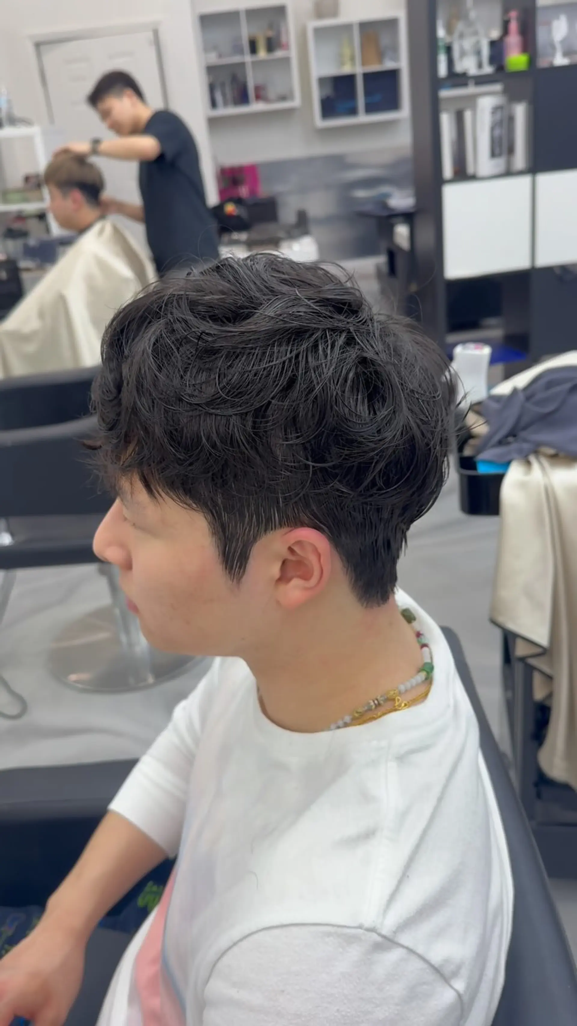 A short men's cut styled with textured wavy perm volume on top and softly tapered sides. 1