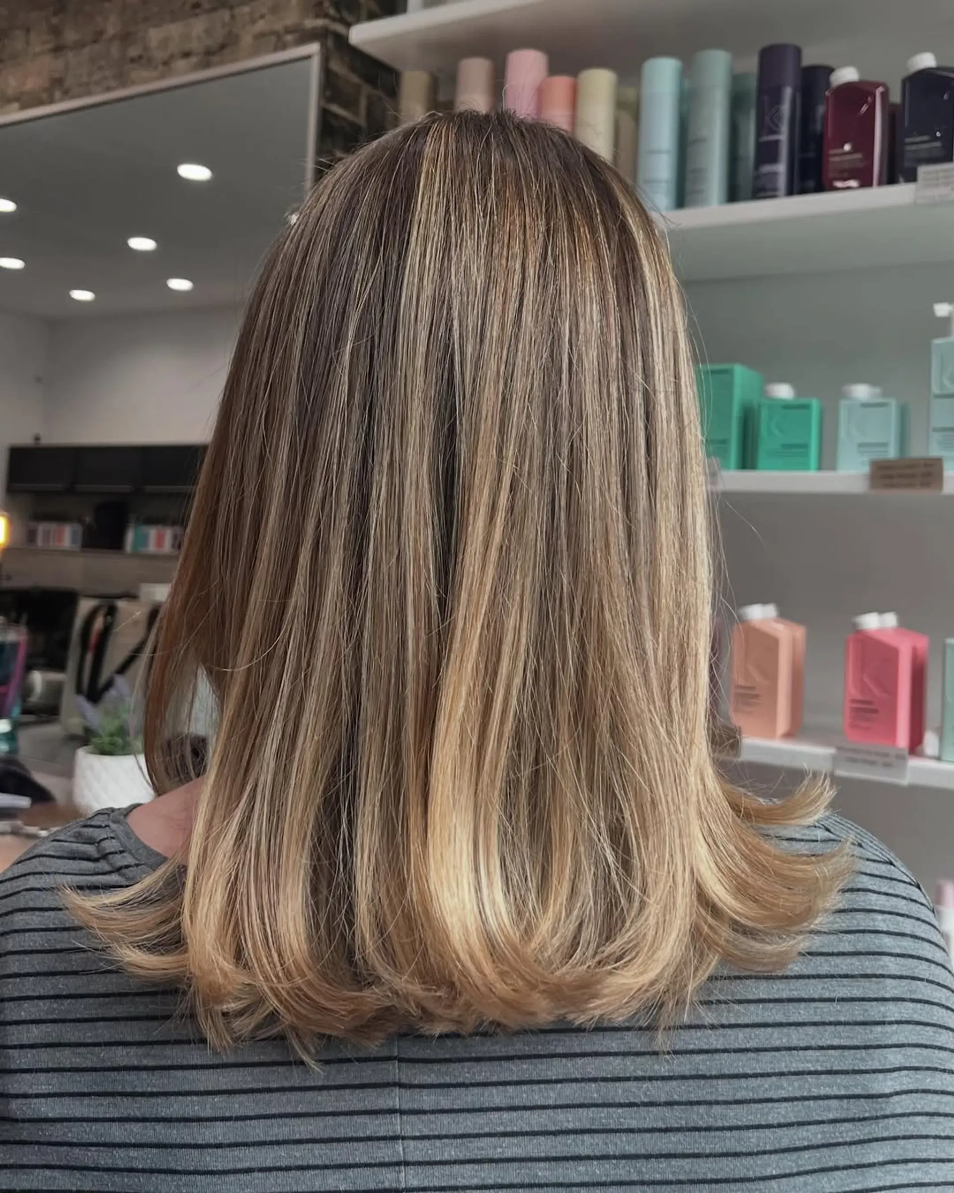 A sleek, shoulder-length lob haircut complemented by a dimensional bronde balayage, seamlessly transitioning from darker roots to brighter golden blonde ends with subtle flipped-out tips. 1