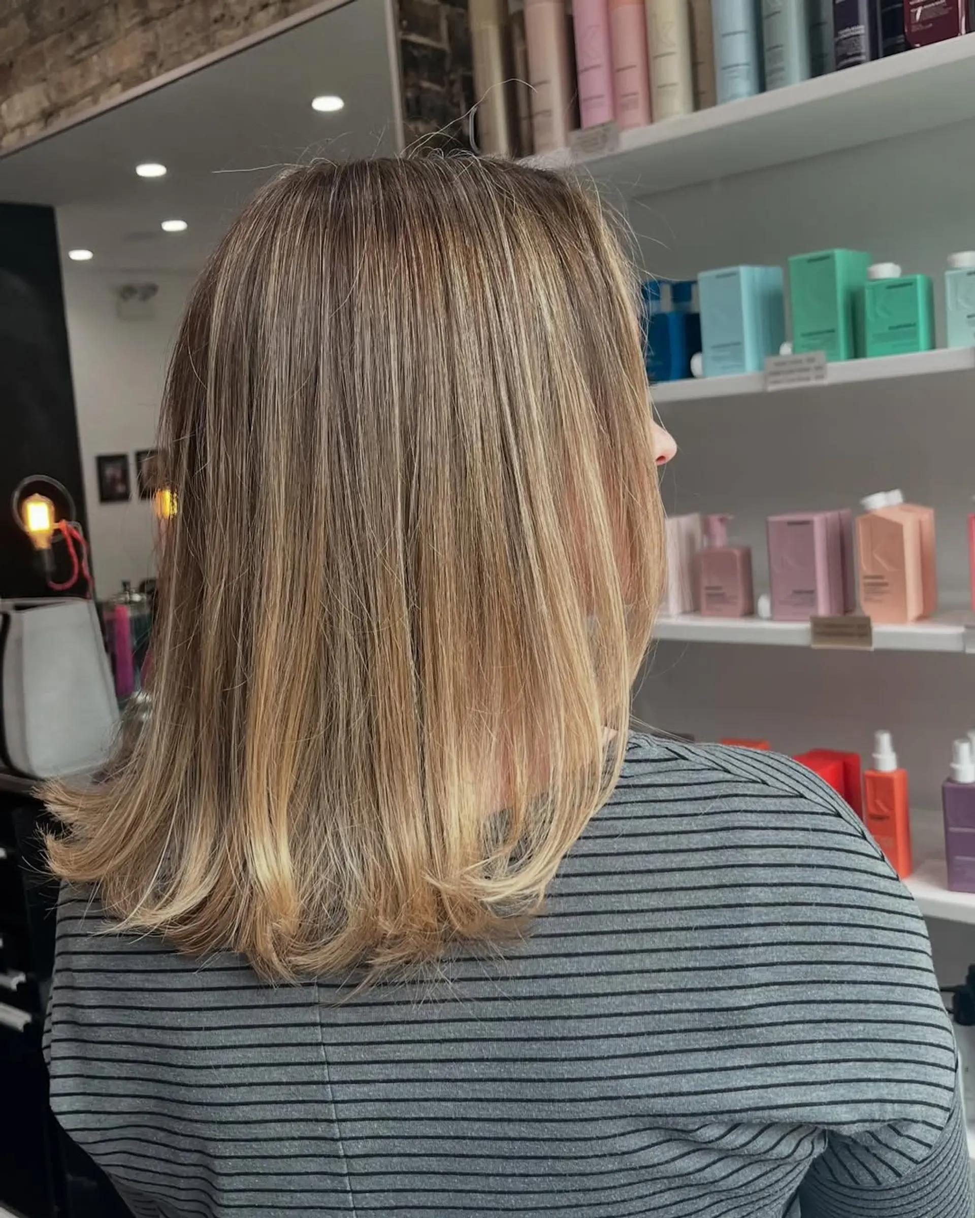 A sleek, shoulder-length lob haircut complemented by a dimensional bronde balayage, seamlessly transitioning from darker roots to brighter golden blonde ends with subtle flipped-out tips. 2