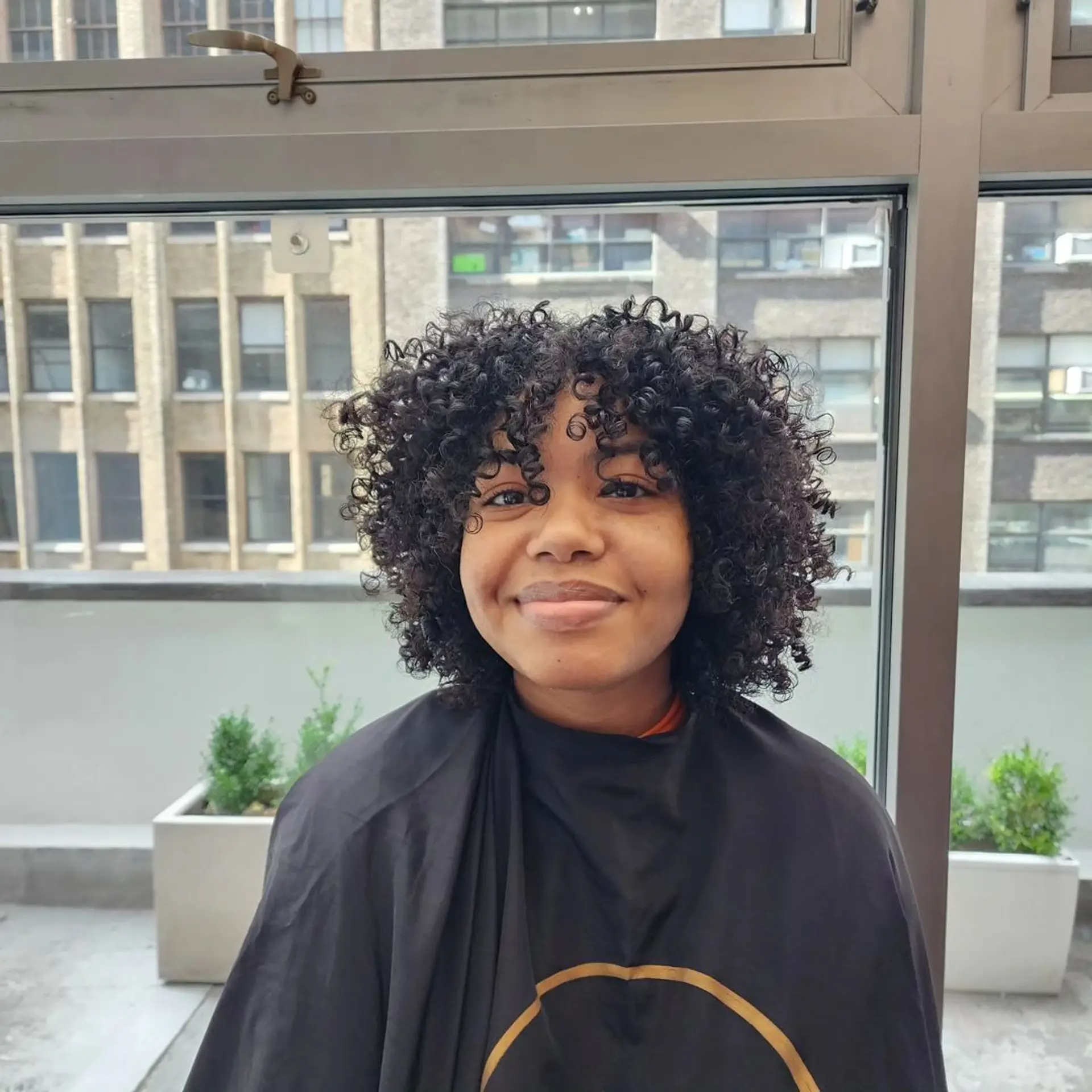 Medium length, naturally curly hair with tight, defined curls and a voluminous appearance. The curls fall around the face, complemented by natural fringe bangs. 1