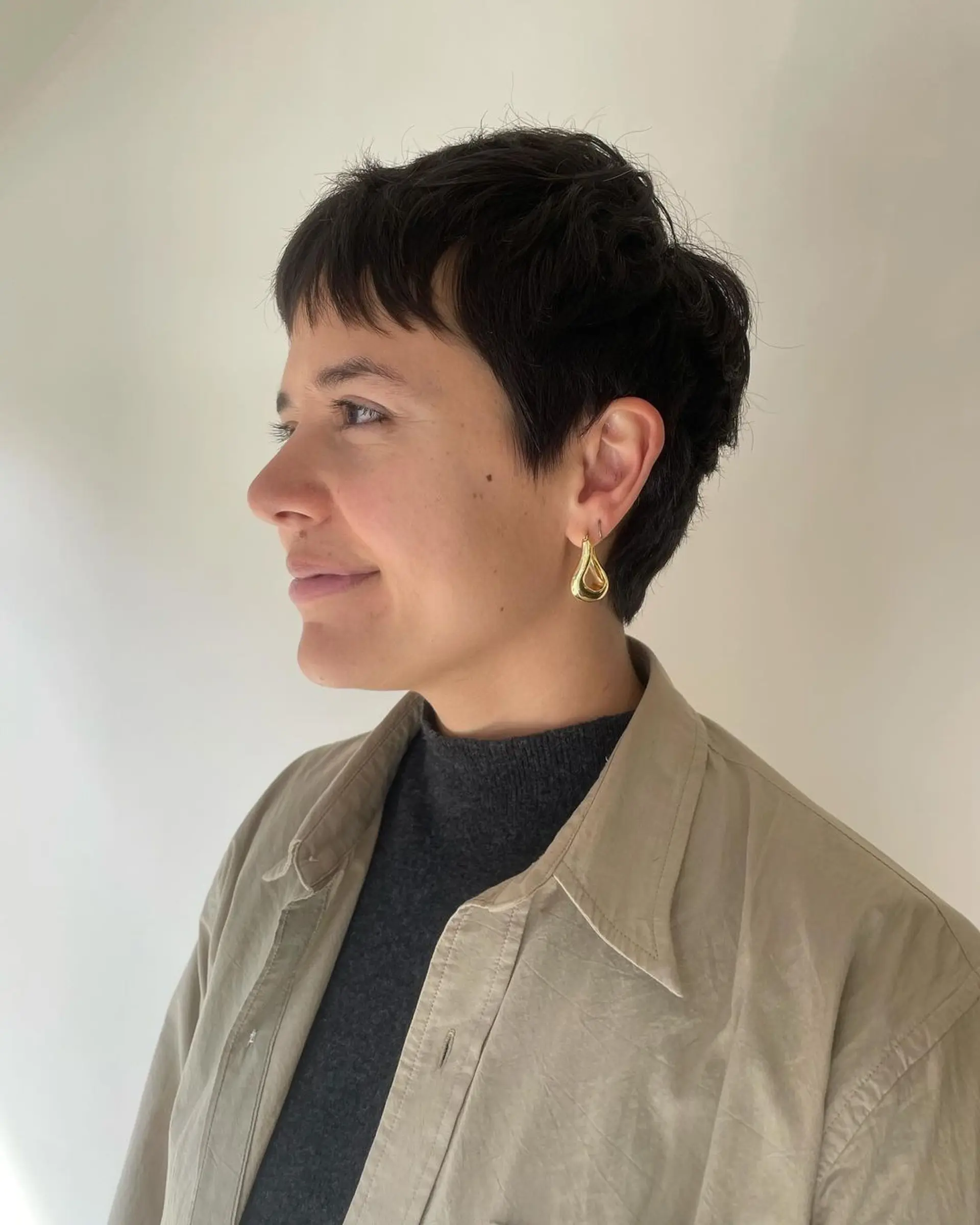 A short, wavy, dark brown pixie cut with textured top and side tapering. Features a subtle fringe framing the face. 1