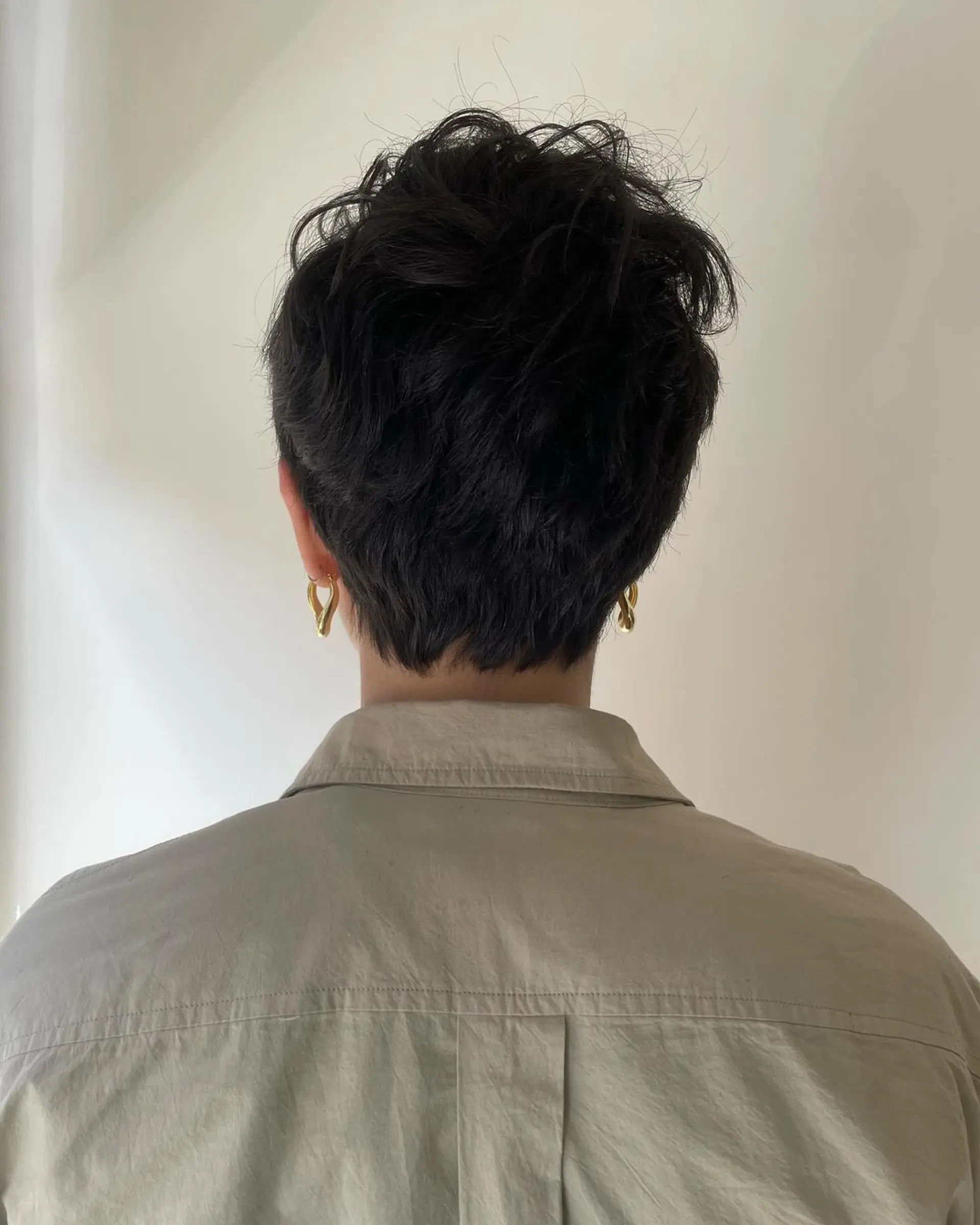 A short, wavy, dark brown pixie cut with textured top and side tapering. Features a subtle fringe framing the face. 2