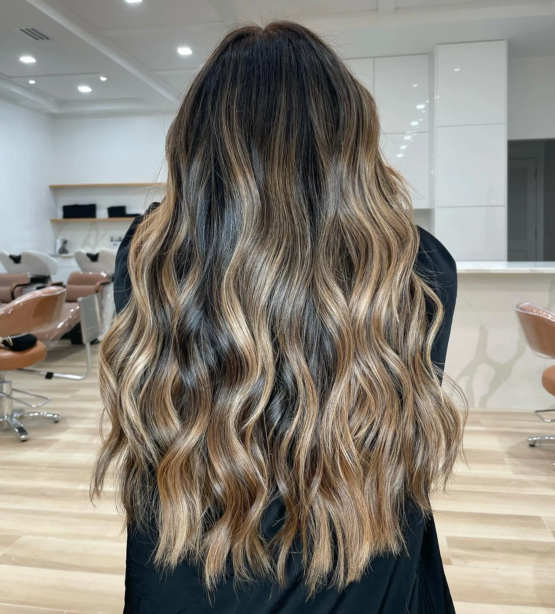 Long cascading hair featuring deep brunette roots blended into dimensional caramel and warm blonde balayage highlights, styled with loose effortless waves. 1