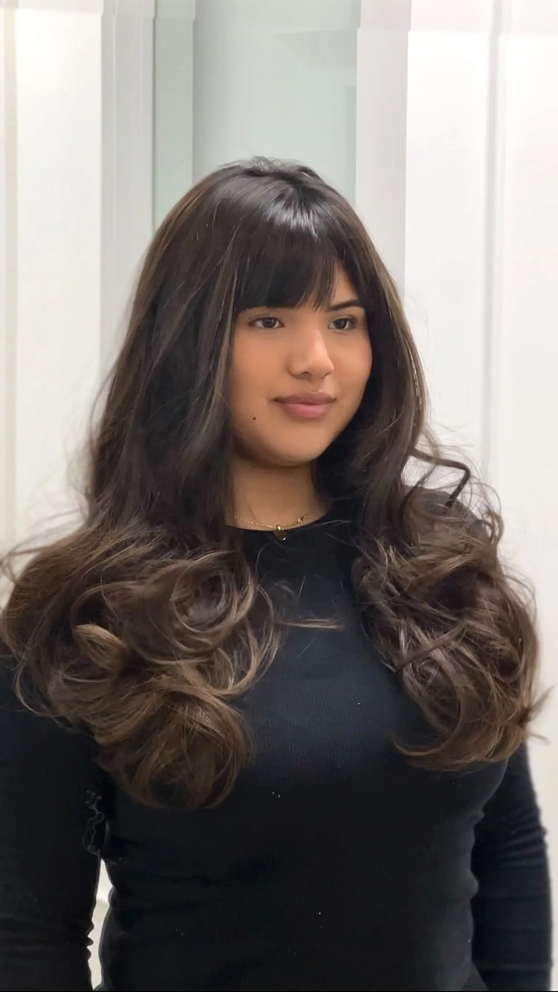 Long layered dark brunette hair with soft warm balayage highlights, styled into bouncy, voluminous curls and paired with eyebrow-grazing full fringe bangs. 1