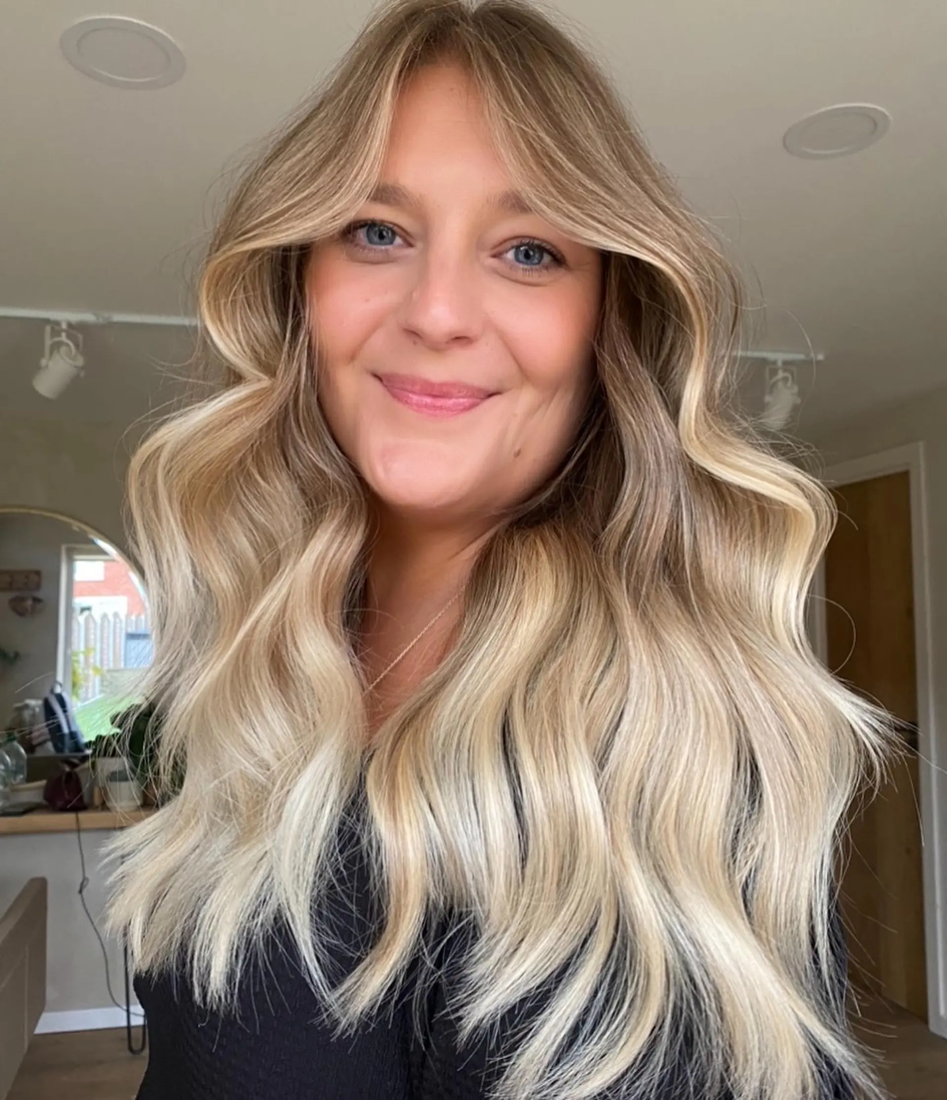 Long dimensional blonde hair styled in soft, voluminous waves with face-framing curtain bangs. 1
