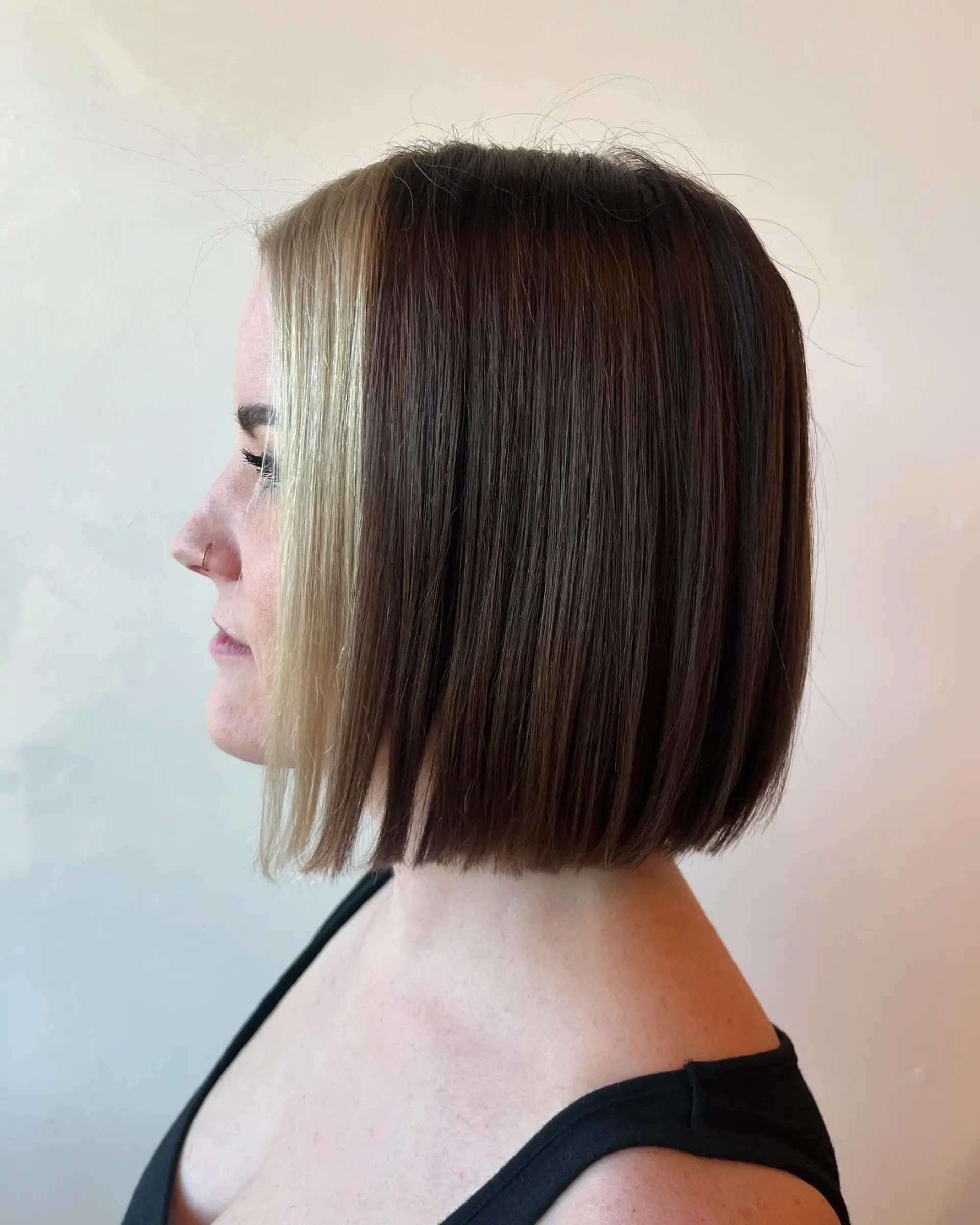 A sleek, chin-length blunt bob featuring deep brunette hair accented by a bold, contrasting blonde money piece framing the face. 1