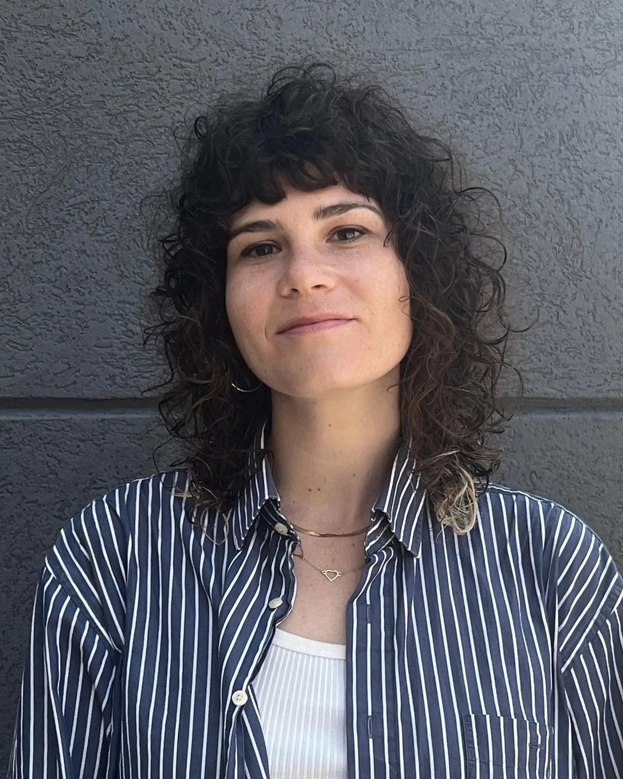 Medium length, curly shag cut with natural voluminous ringlets, a wispy fringe, and subtle blonde highlights over a dark brown base, creating a soft, tousled look with textured ends.